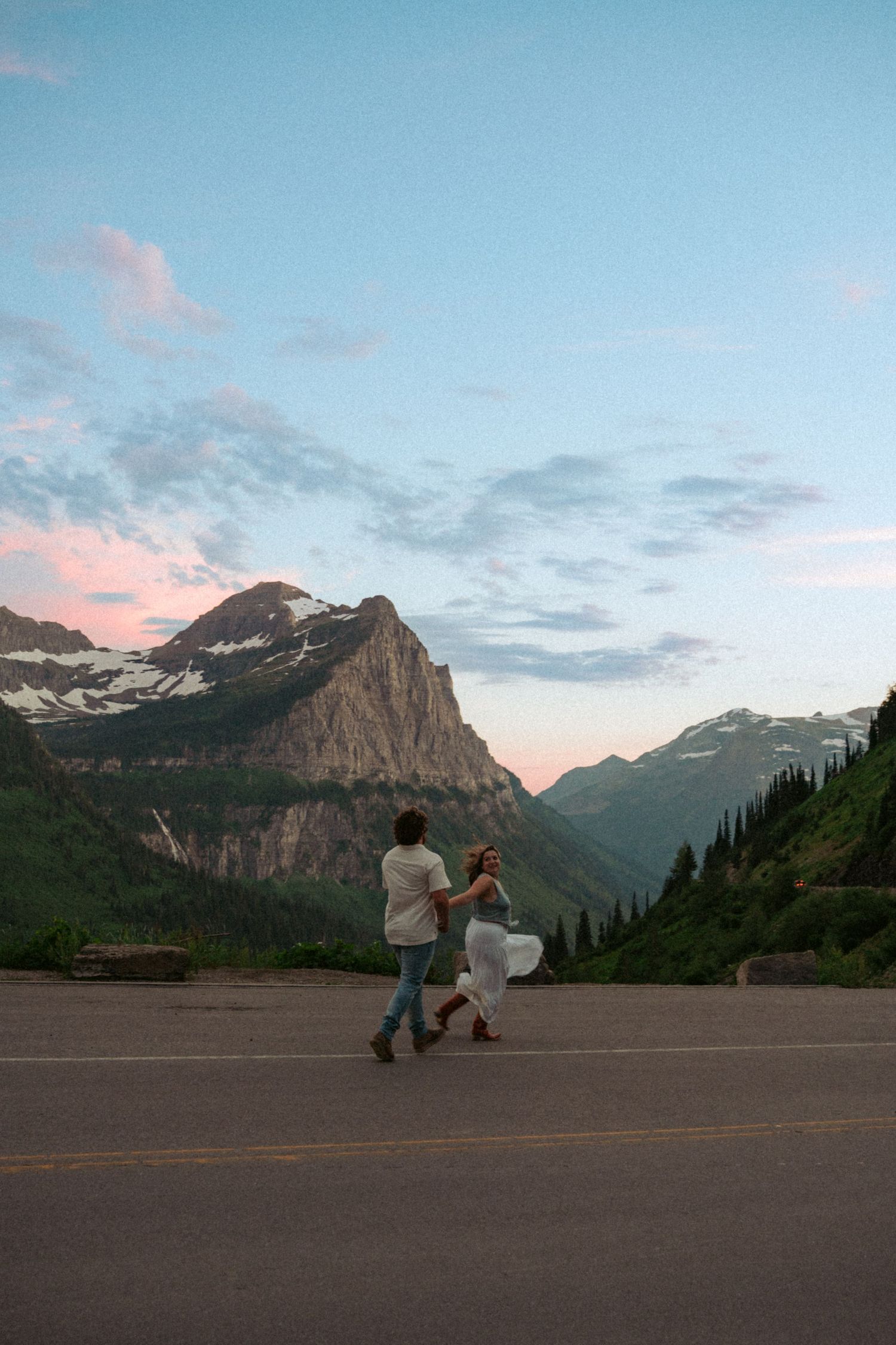 Couple running hand-in-hand on a mountain road at sunset with snow-capped peaks and lush green valleys in the background.