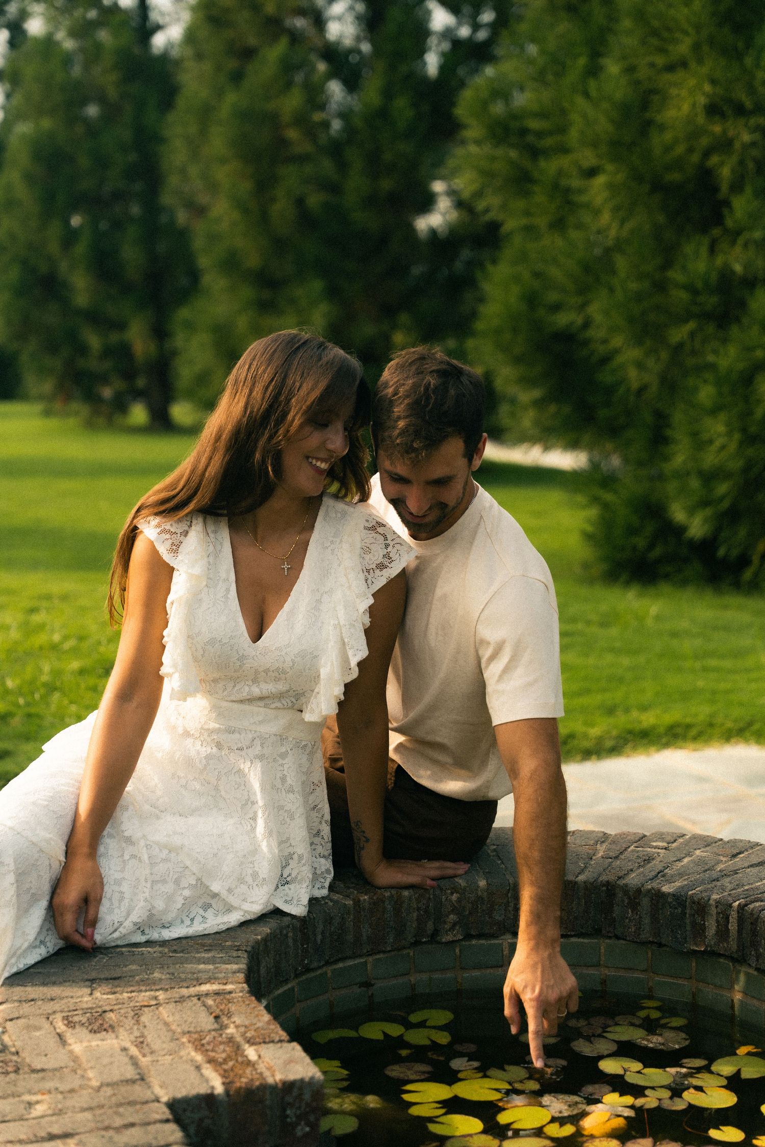 A couple in white clothing leaning over a stone wall with lily pads in the water below.