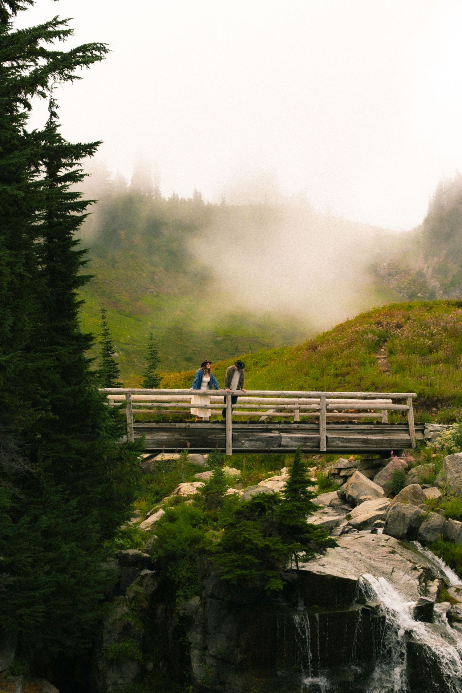 Couple stands on wooden bridge over waterfall with misty mountain landscape in background.