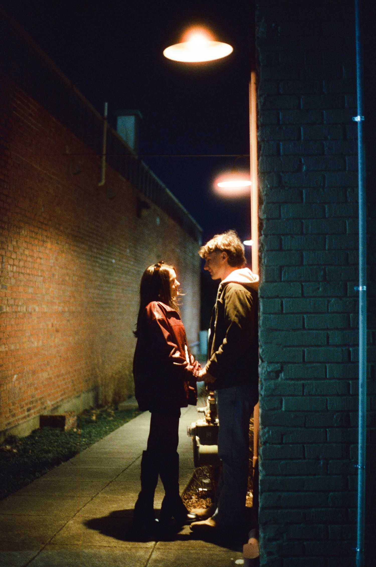 Two people stand in a dimly lit alleyway against a brick wall under a hanging lamp at night.