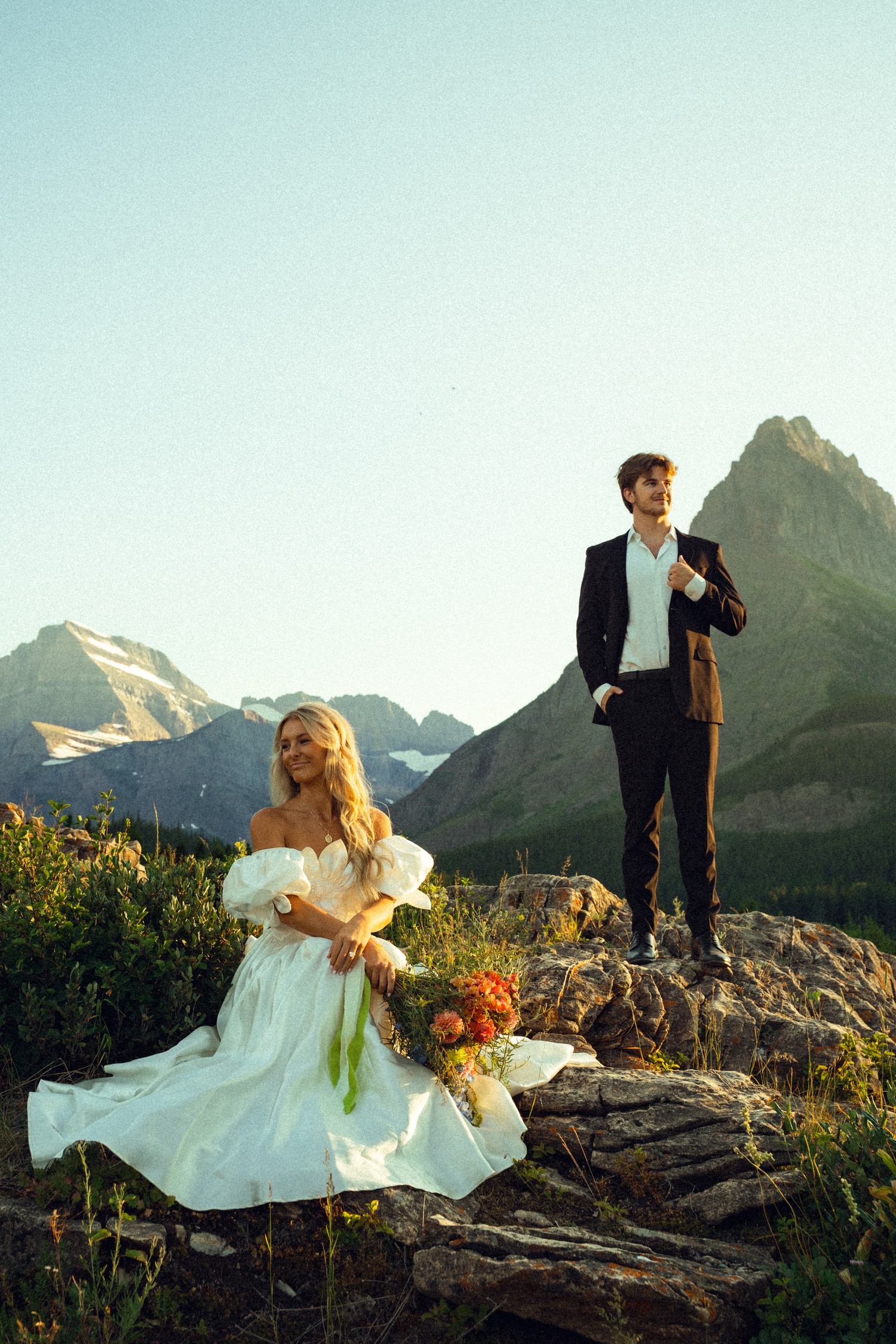 Bride in white gown sits on rocks as groom stands behind her with stunning mountain landscape at golden hour.