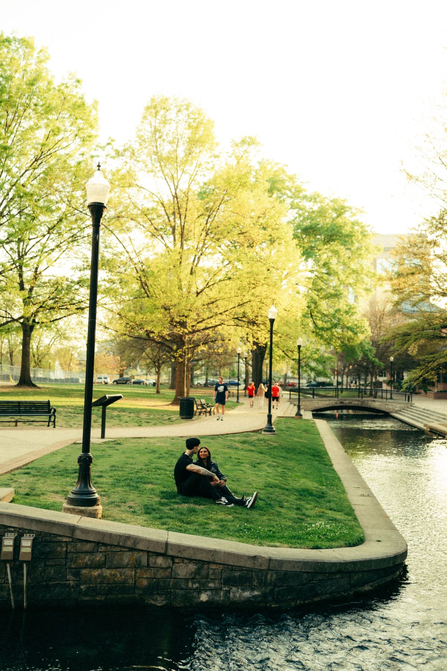Couple sitting on grassy canal bank in a scenic urban park with tall trees, lamp posts, and a bridge on a sunny day.