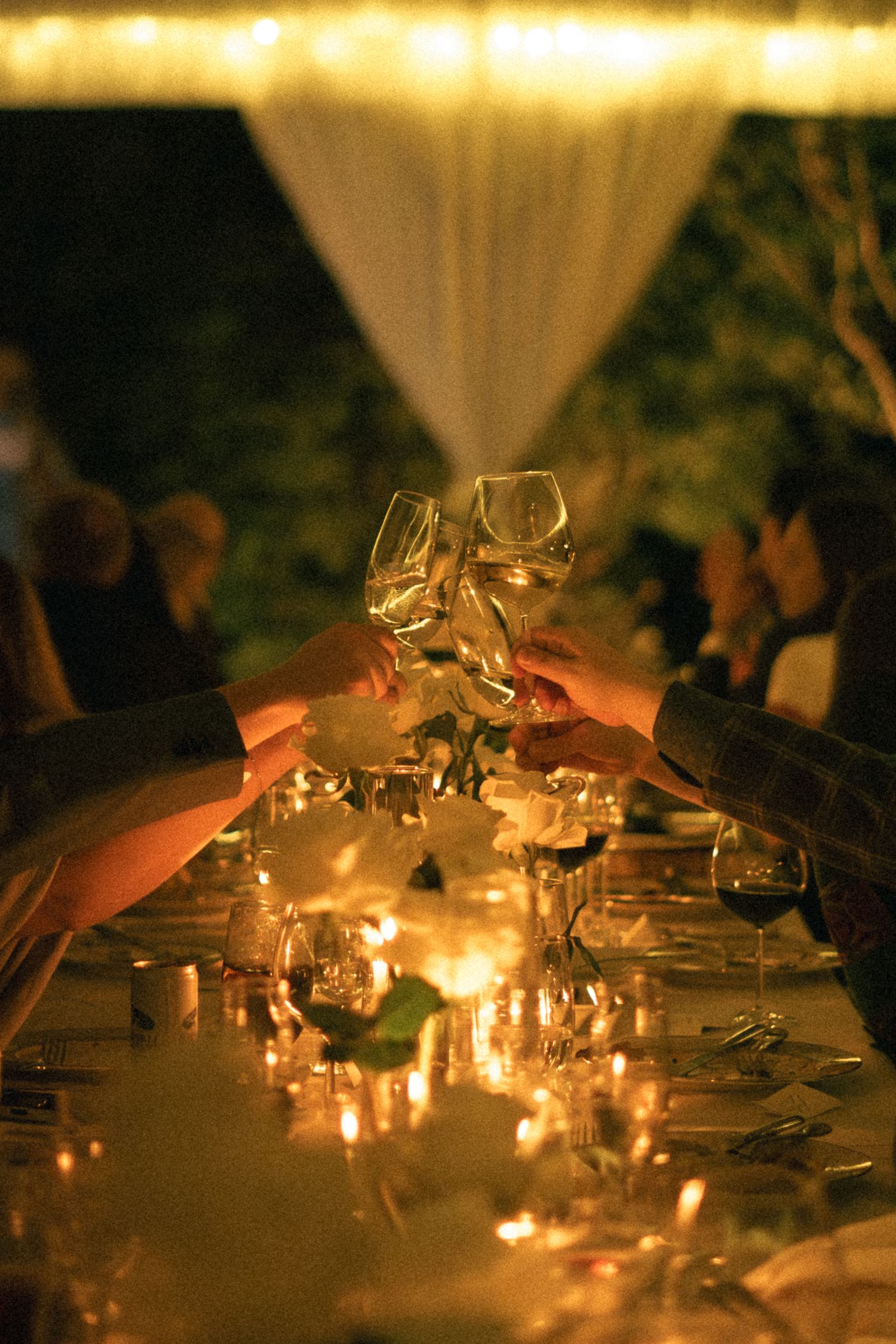 Wine glasses clink together in a candlelit toast during an evening celebration.