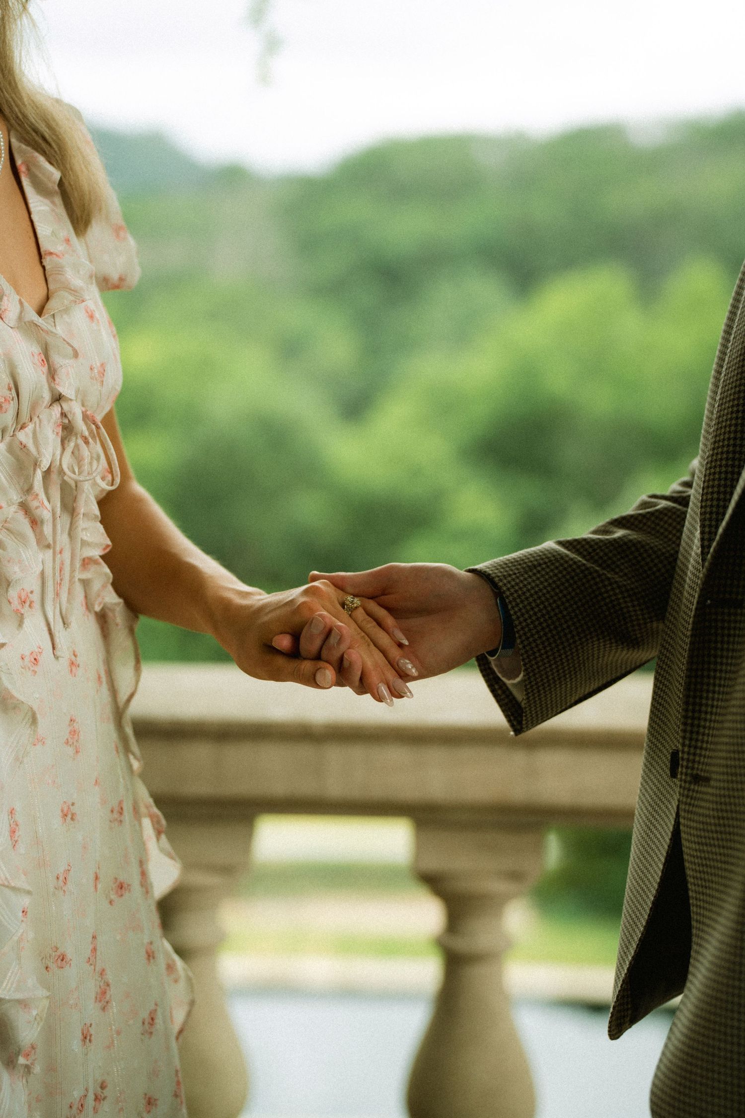 Couple holding hands on a balcony with lush green trees in the background, wearing floral dress and plaid suit.