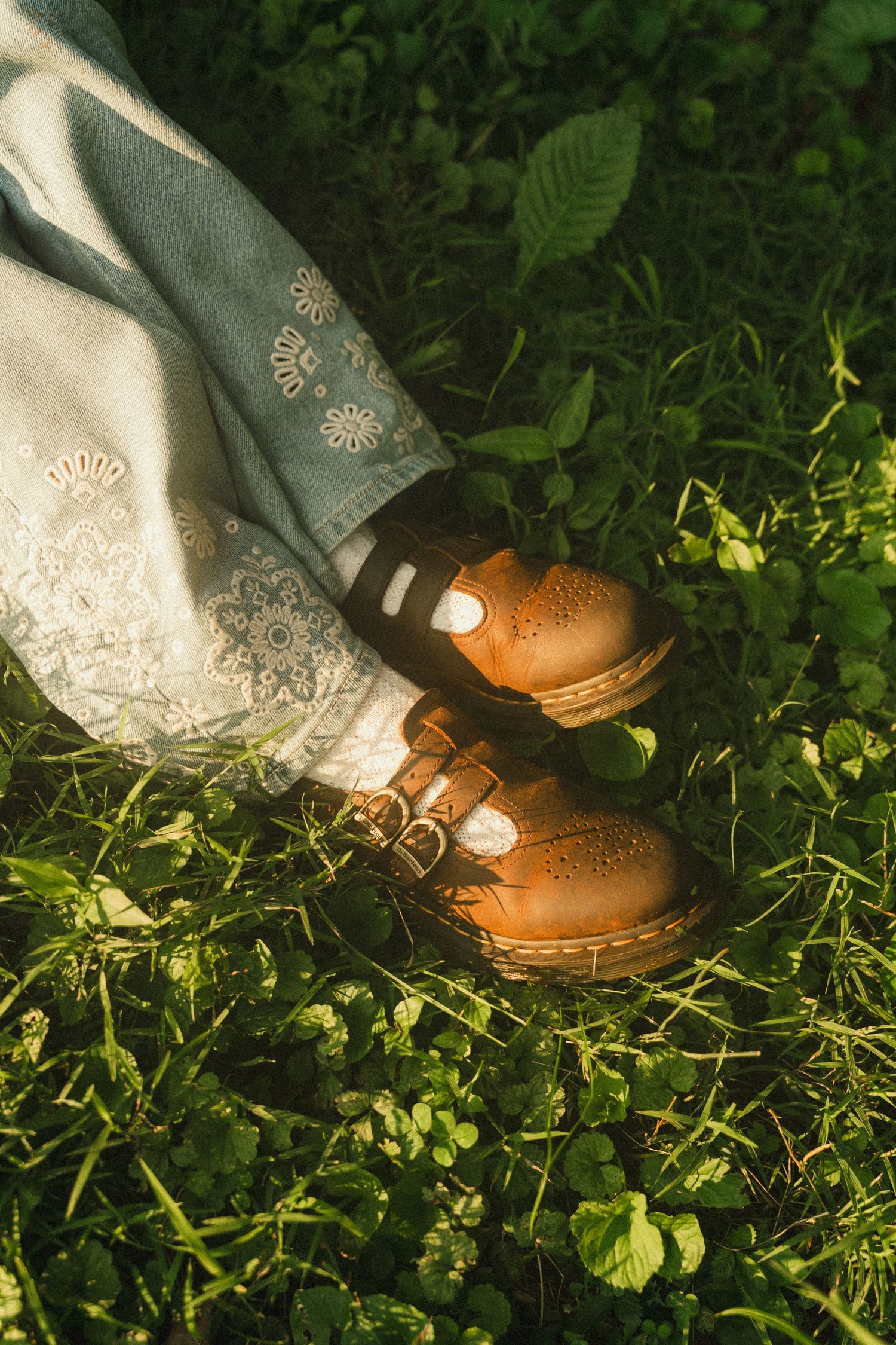 Person wearing tan leather Dr. Martens brogues and a light blue embroidered skirt standing on lush green grass.