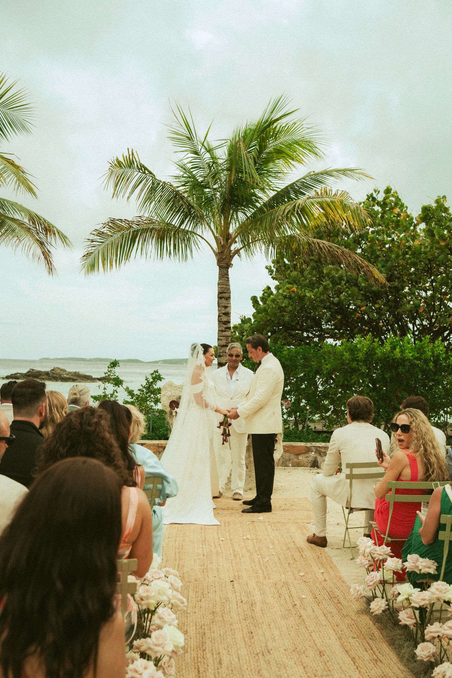 Outdoor beach wedding ceremony with couple exchanging vows under a palm tree, flanked by white floral arrangements and seated guests.
