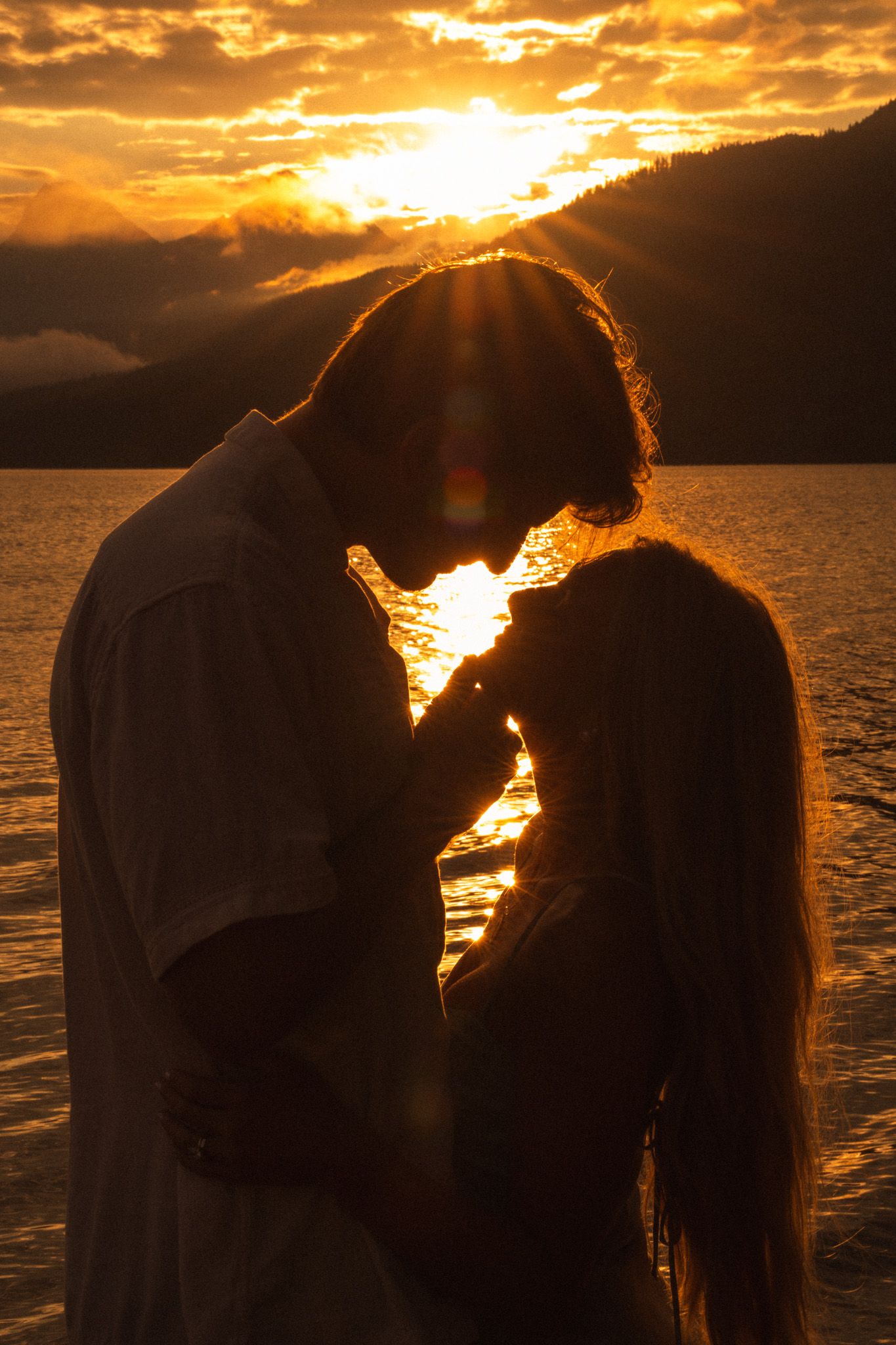 Romantic couple silhouetted against a golden sunset over a lake with mountains, sharing an intimate moment.