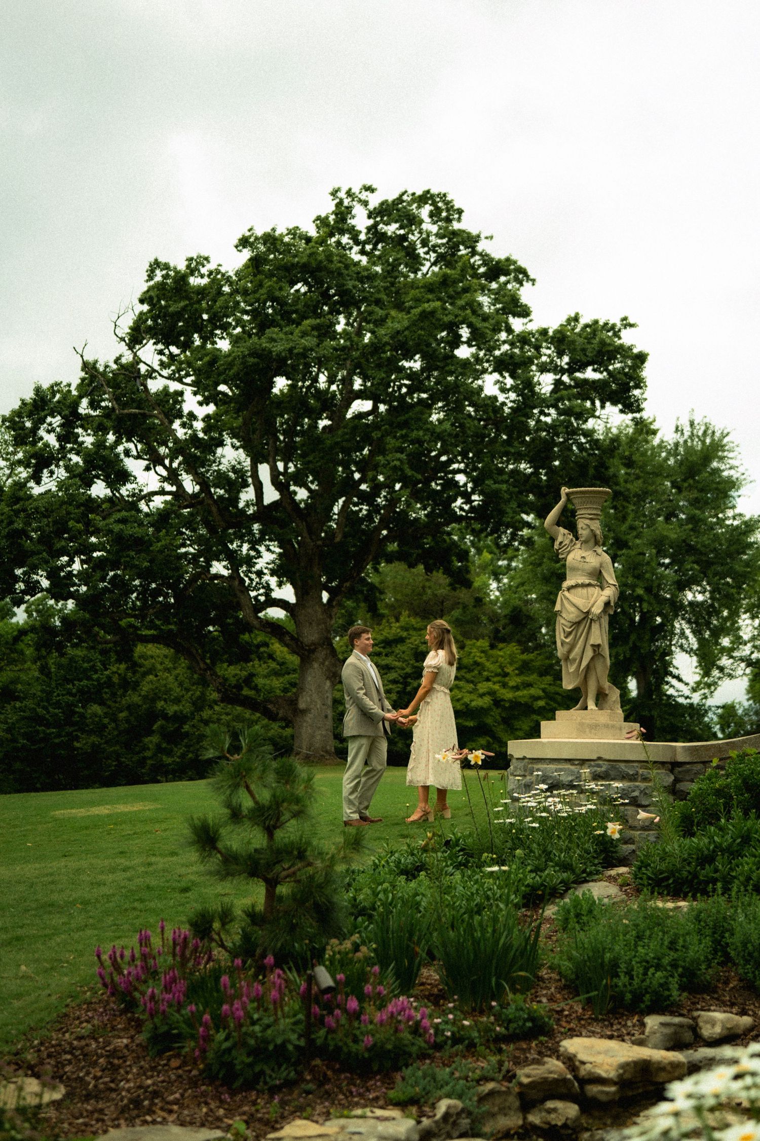 Couple holding hands in a lush garden beside a classical stone statue, surrounded by colorful flowers and trees.