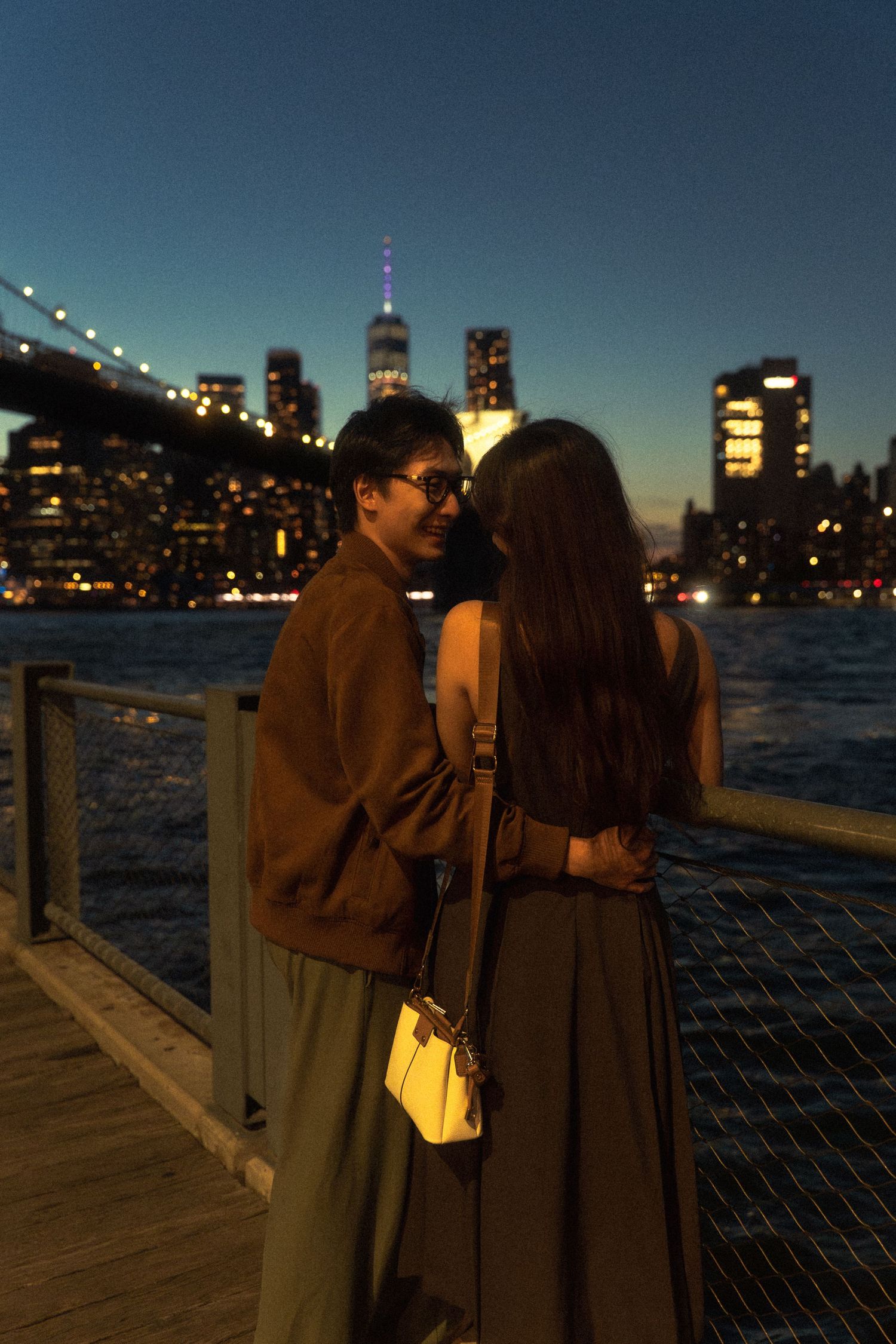 A romantic couple embraces at dusk on a waterfront boardwalk with illuminated city skyline in the background.