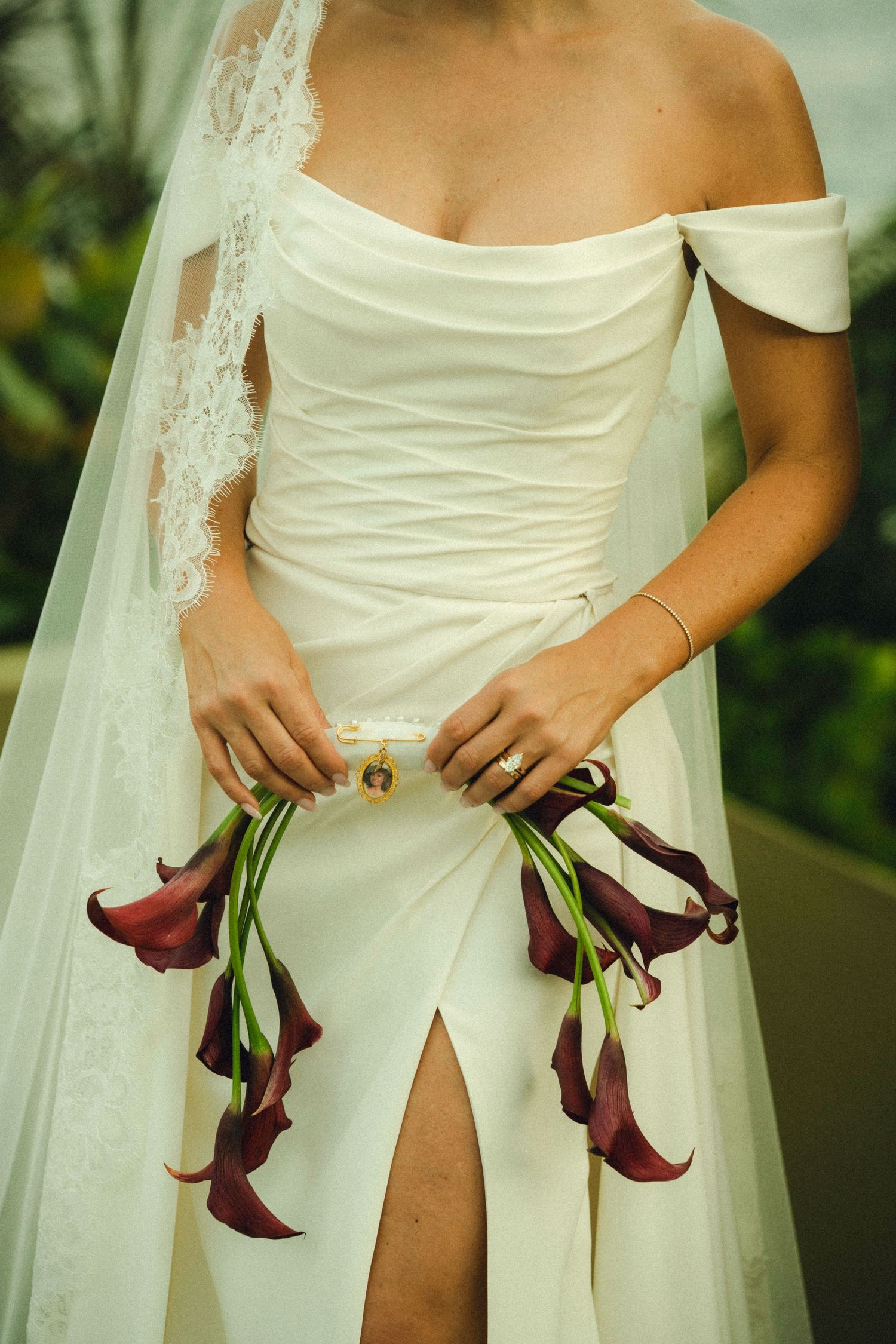 Bride in off-shoulder white gown holding burgundy calla lily bouquet with lace veil, elegant wedding day look.