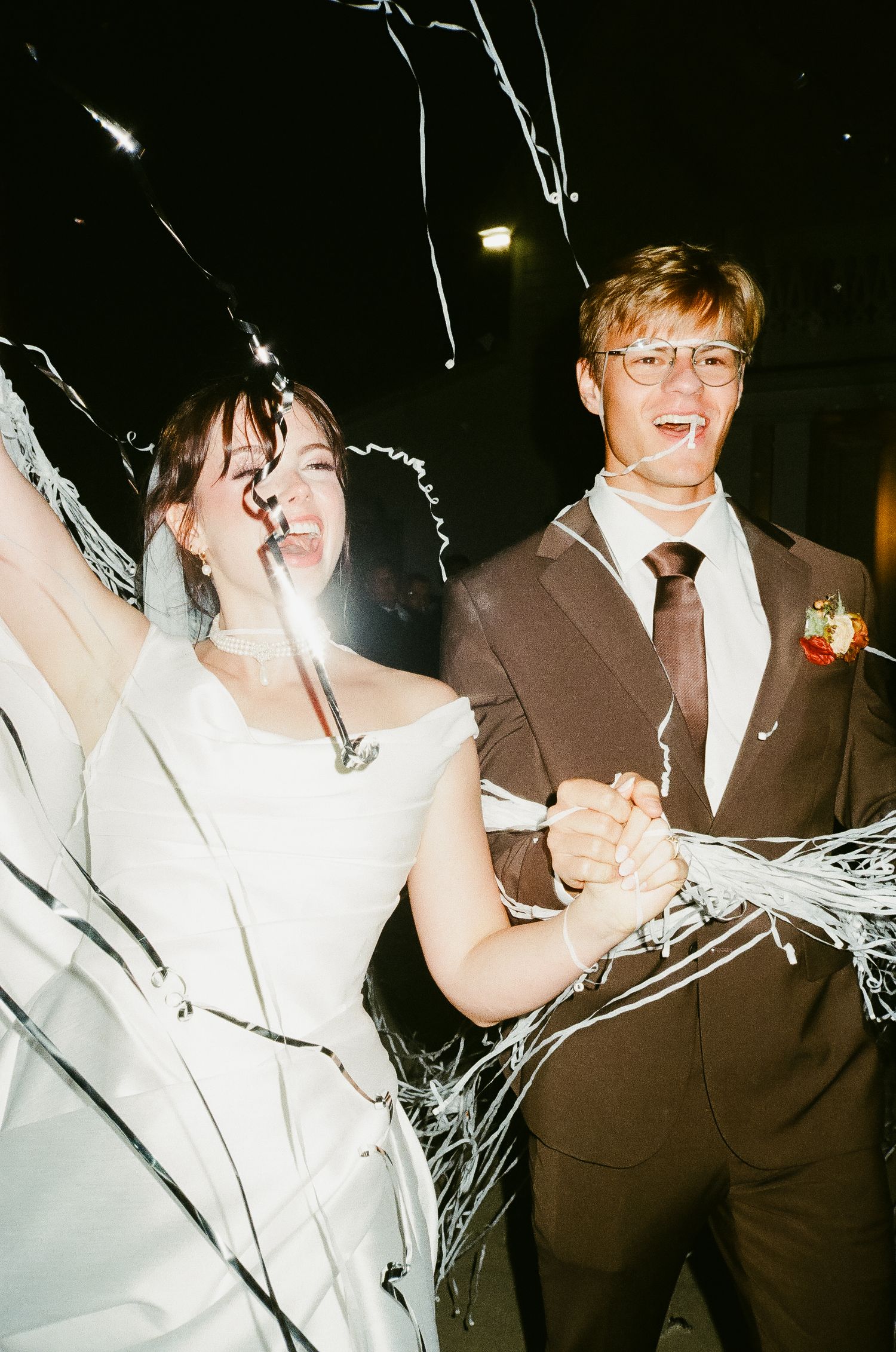 Joyful newlyweds celebrate with sparklers and streamers, bride in white gown and groom in brown suit, wedding exit.