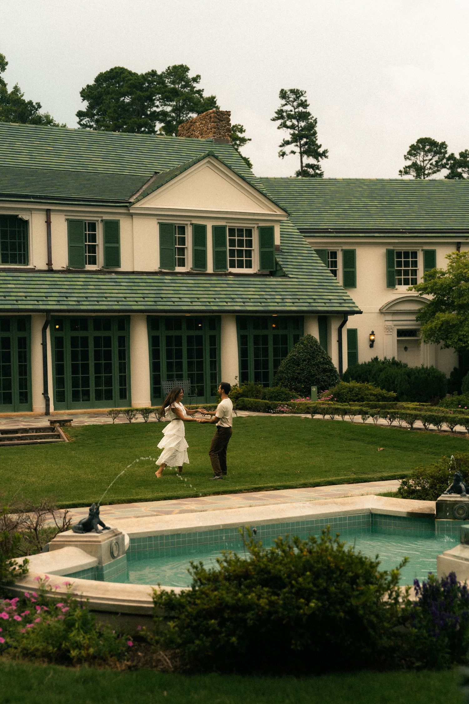 A grand white house with a swimming pool and manicured lawn on a cloudy day.