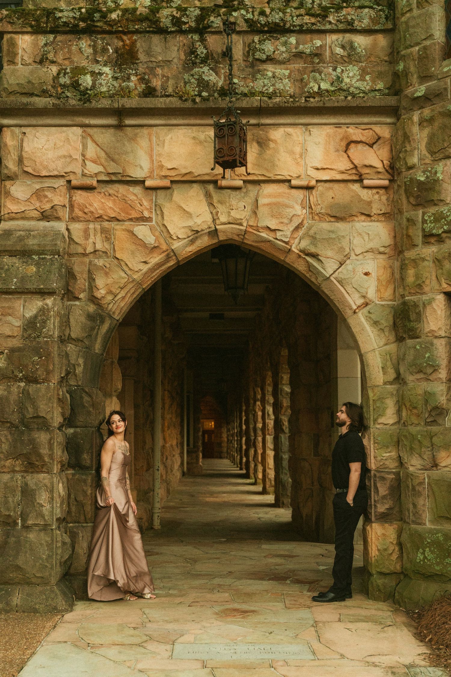 Couple posing under stone archway corridor of historic castle building, woman in elegant gown, man in black attire.