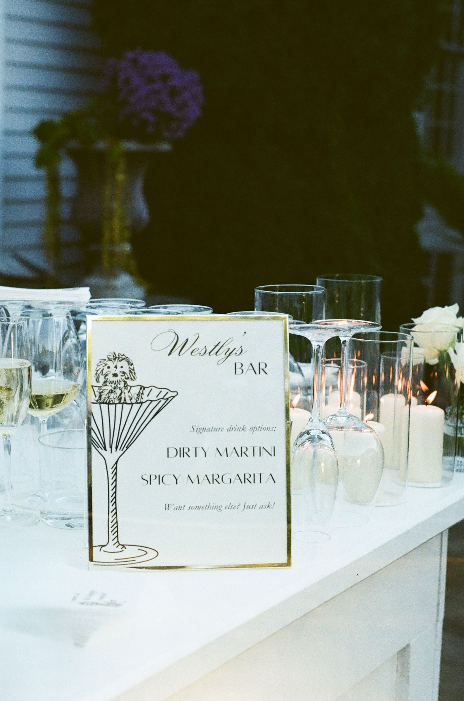 Wedding bar menu sign displaying cocktail options next to empty glasses on white table.