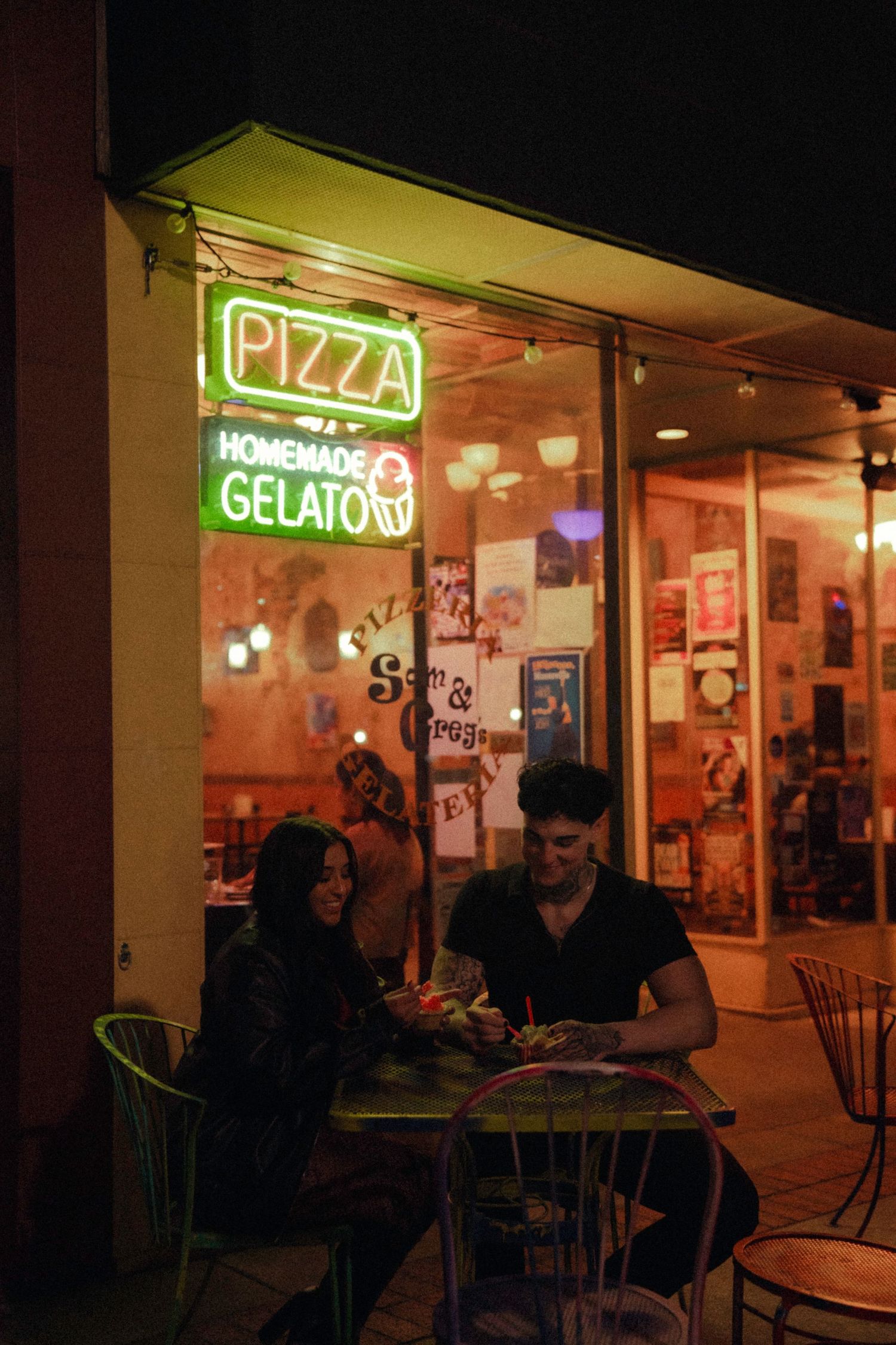 Couple dining outside a pizza and homemade gelato restaurant at night, illuminated by neon signs.