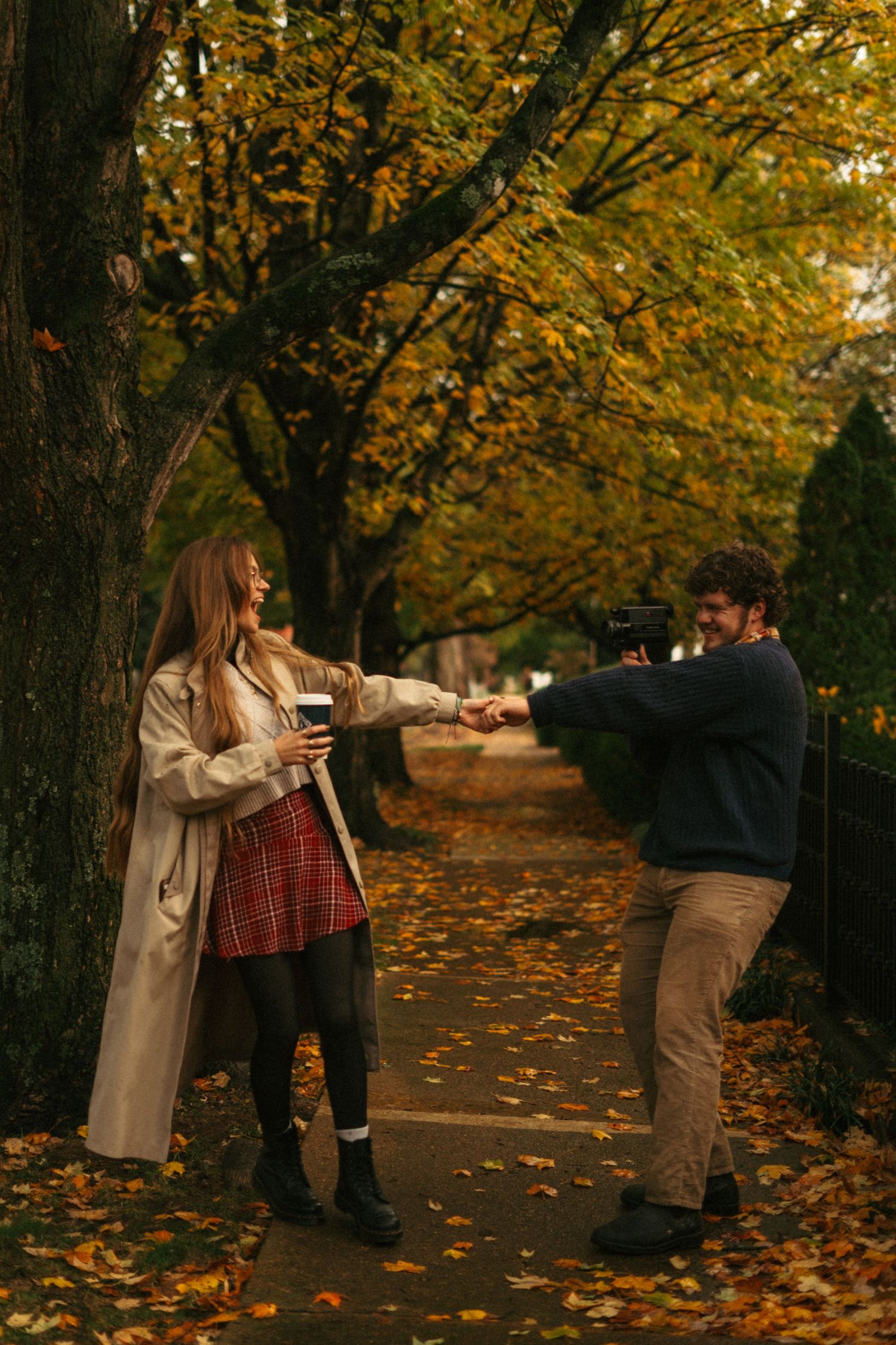 A couple playfully interacting among fallen autumn leaves on a tree-lined path.