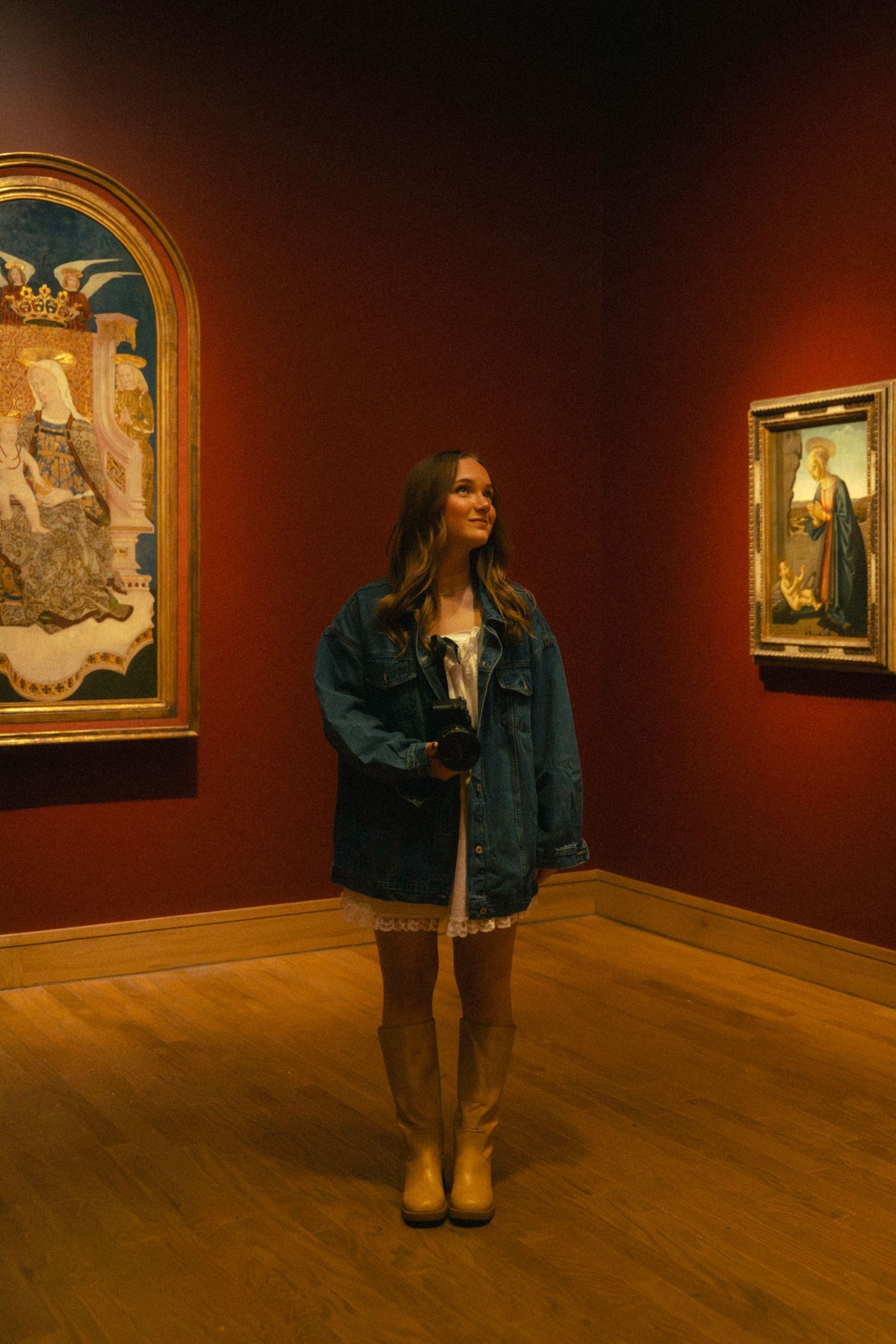Museum visitor in denim jacket and boots stands contemplating classical paintings on deep red gallery walls.
