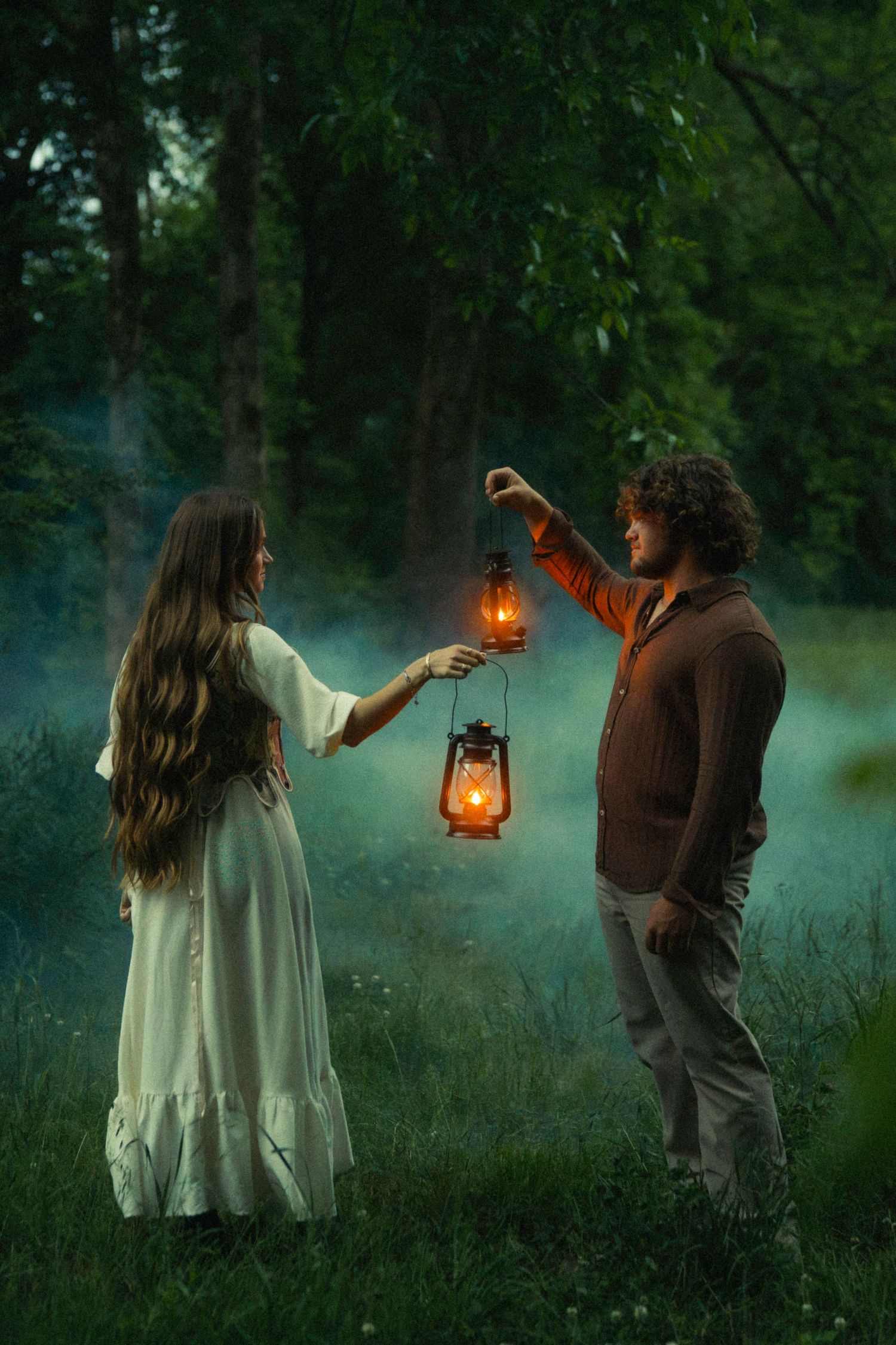 A couple holding glowing lanterns in a misty forest at dusk, woman in white dress, man in brown sweater.