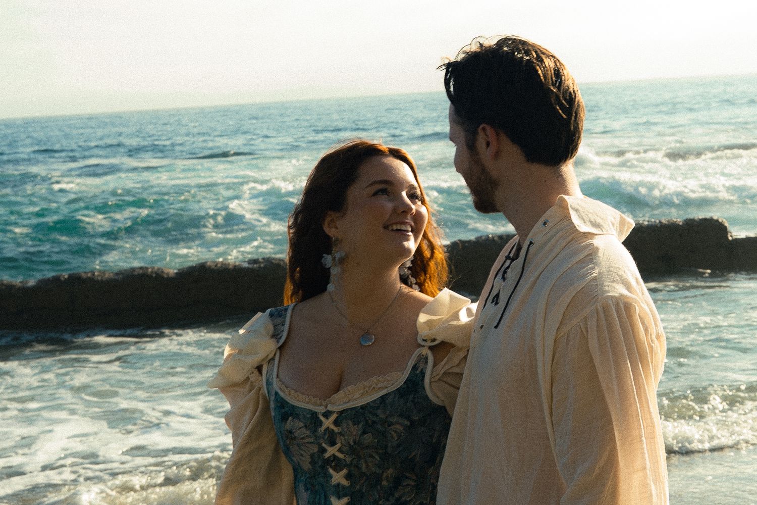 A couple in Renaissance costumes share a romantic moment by the ocean waves on a sunny day.
