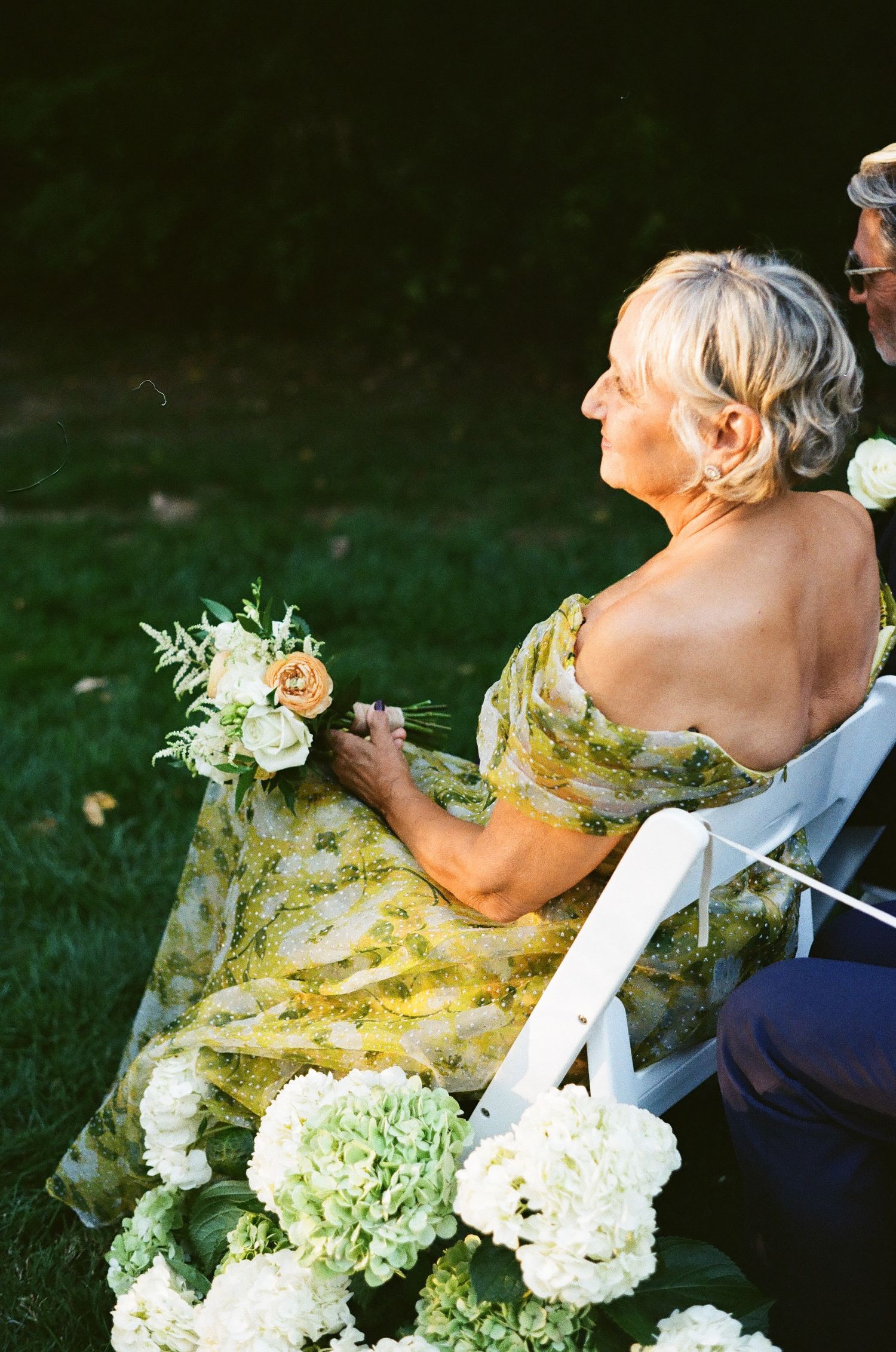 Guest in a floral dress holding flowers at an outdoor wedding ceremony.