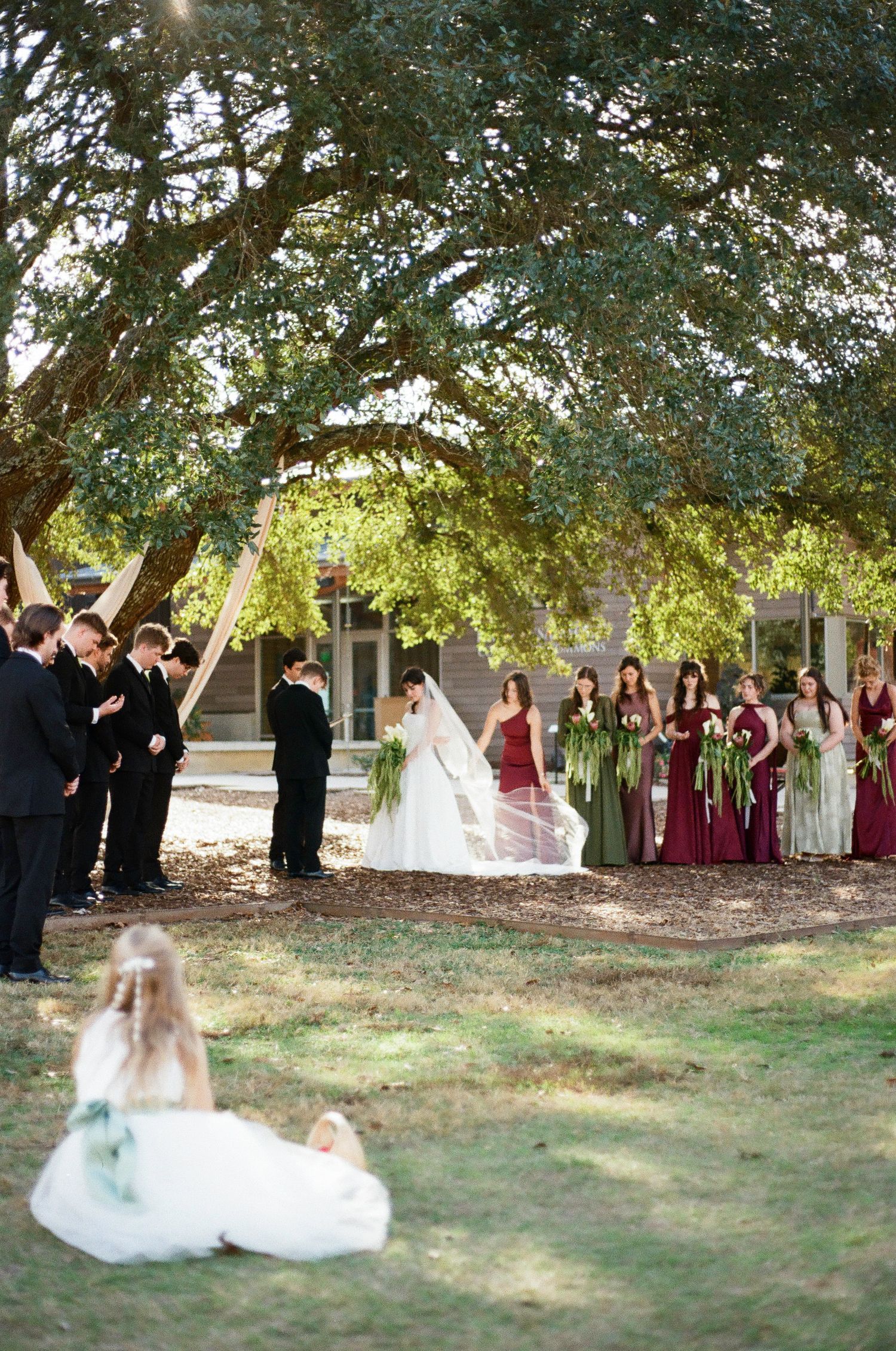 Outdoor wedding ceremony under trees with wedding party and guests gathered.