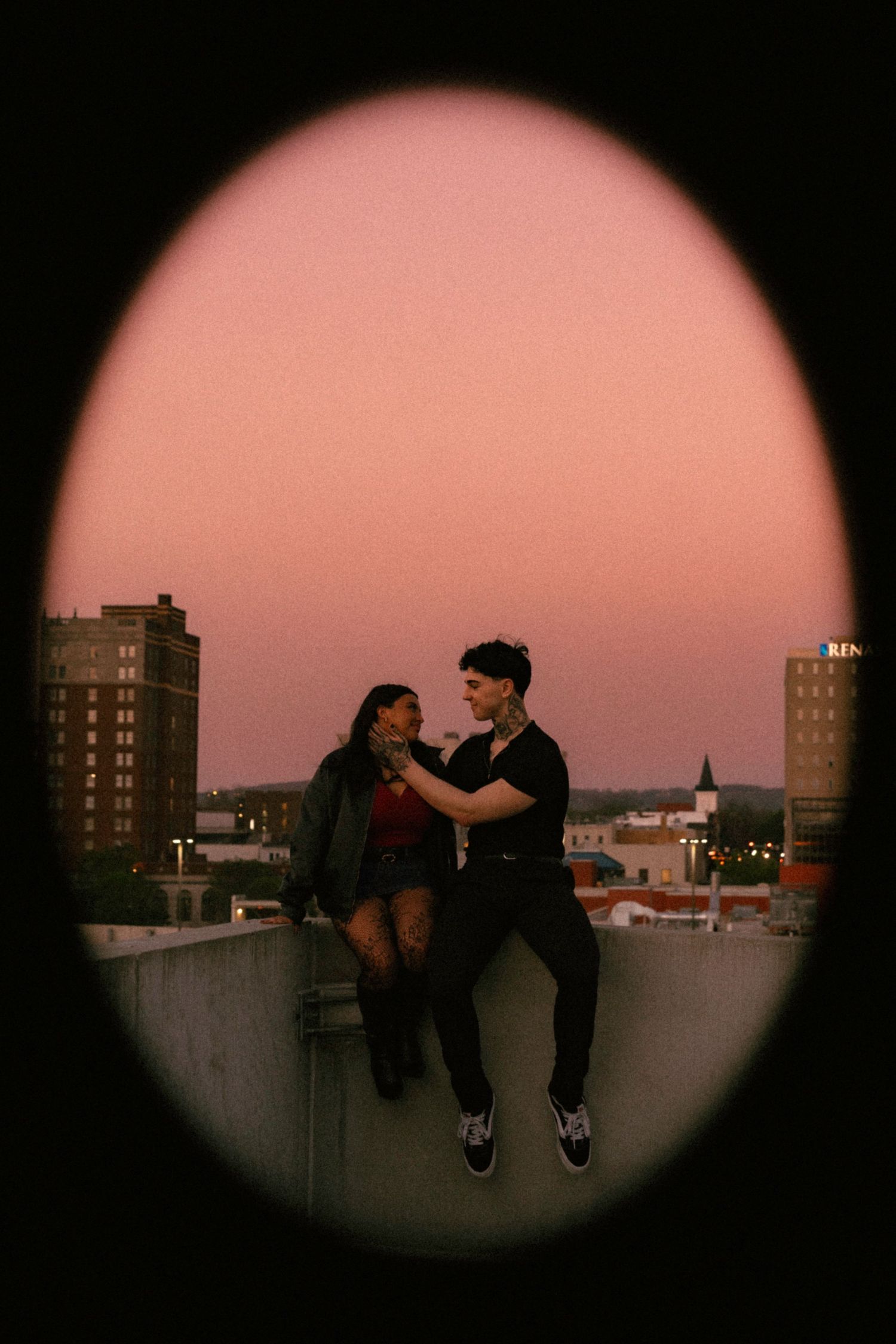 Couple sitting on rooftop edge at sunset with pink sky and city skyline, viewed through oval frame.
