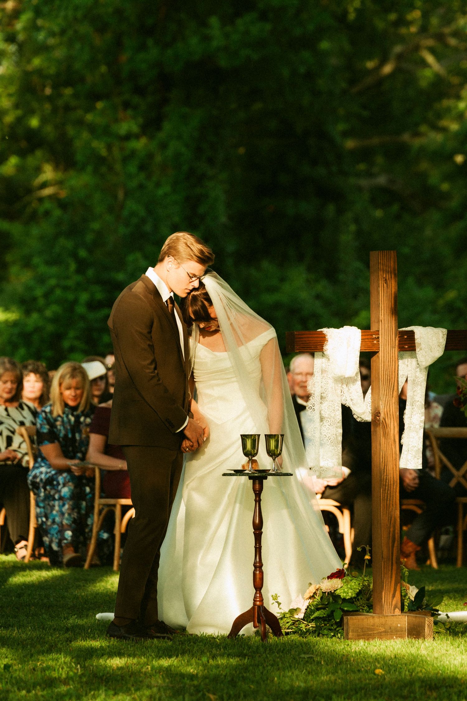 Bride and groom bow heads in prayer at outdoor wedding ceremony beside wooden cross and communion chalices.