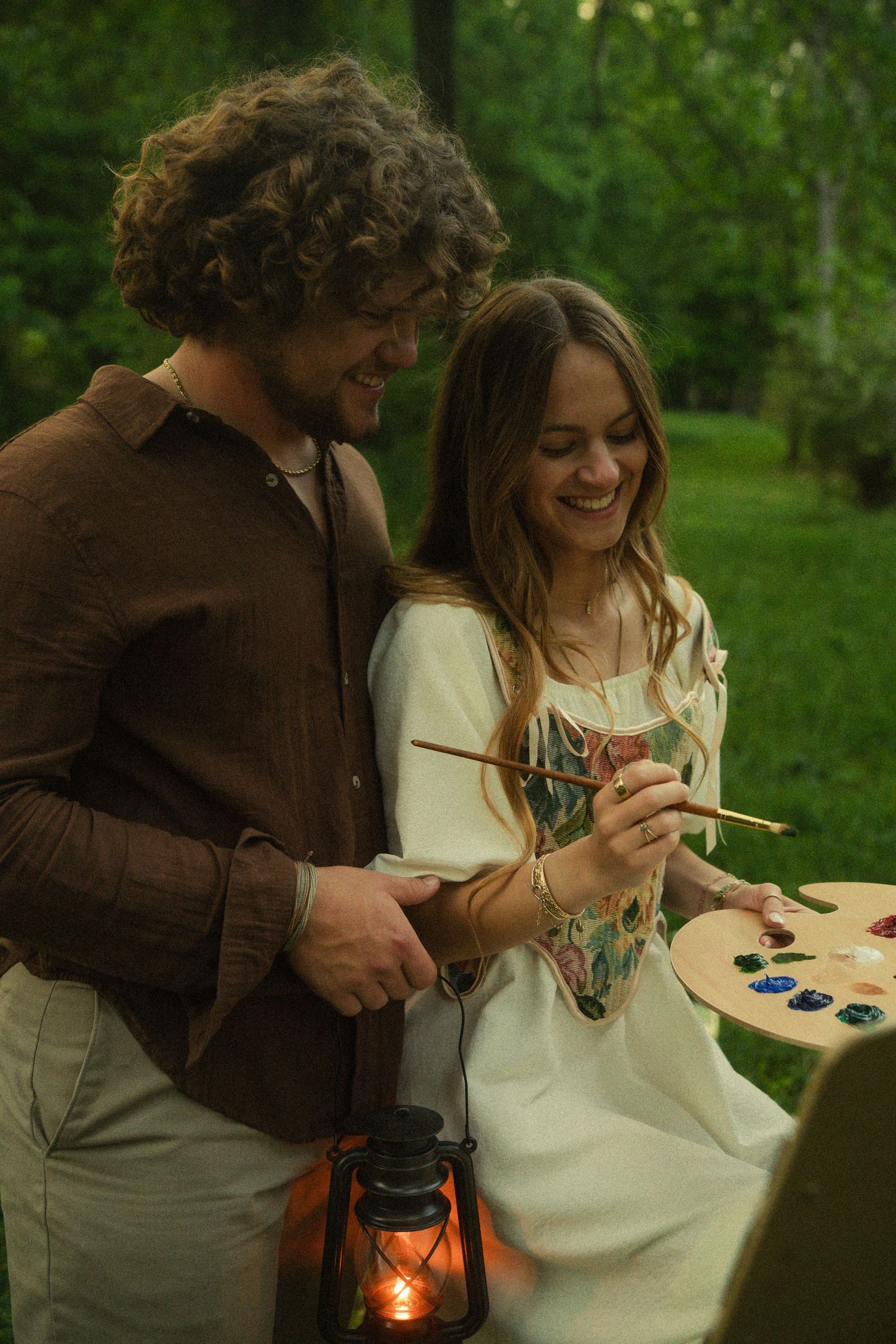 A smiling couple painting outdoors in a green park, woman holds palette and brushes, man carries a lantern.
