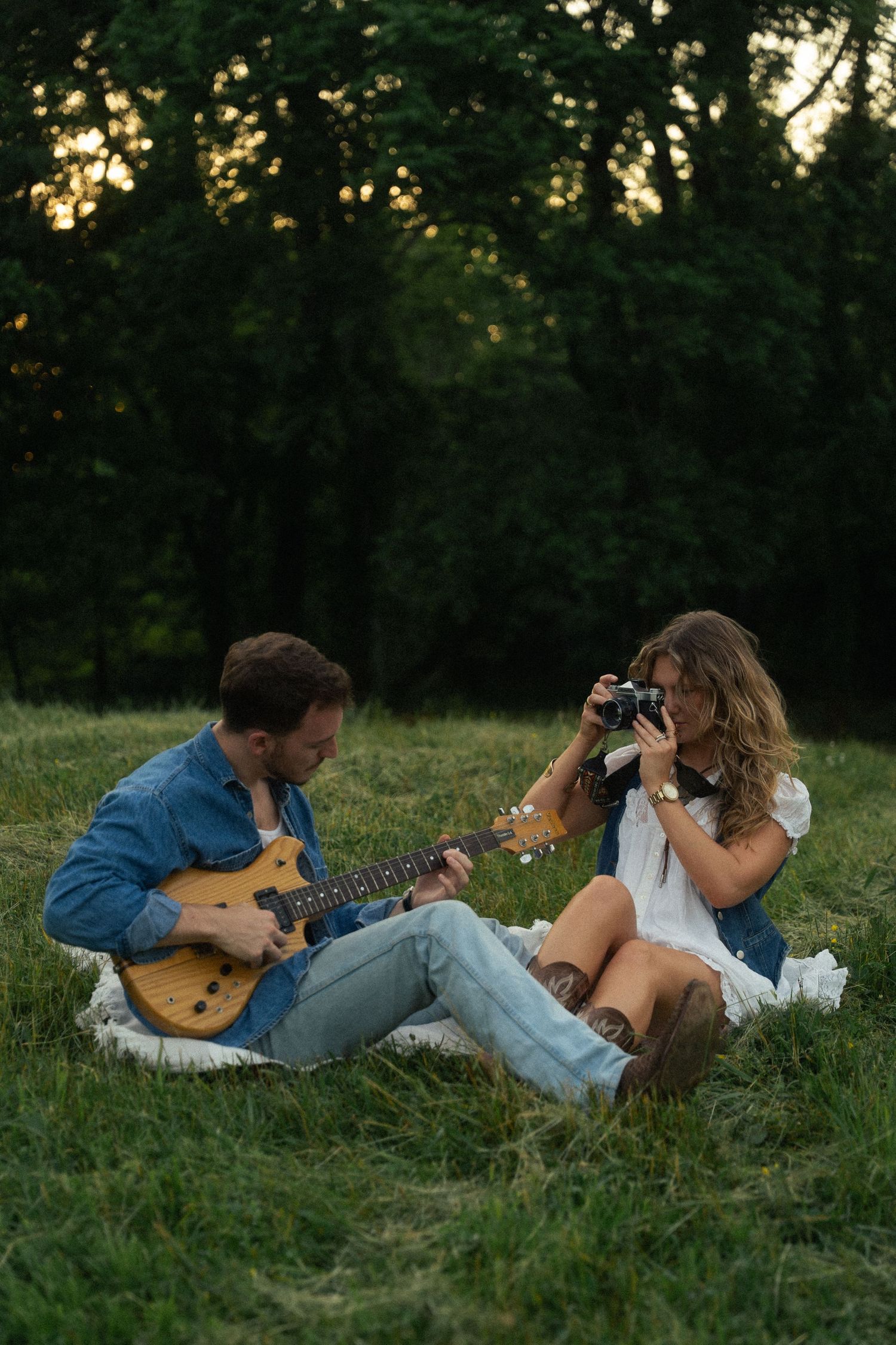 Two people relax on grass while one plays guitar and another takes photos at dusk.
