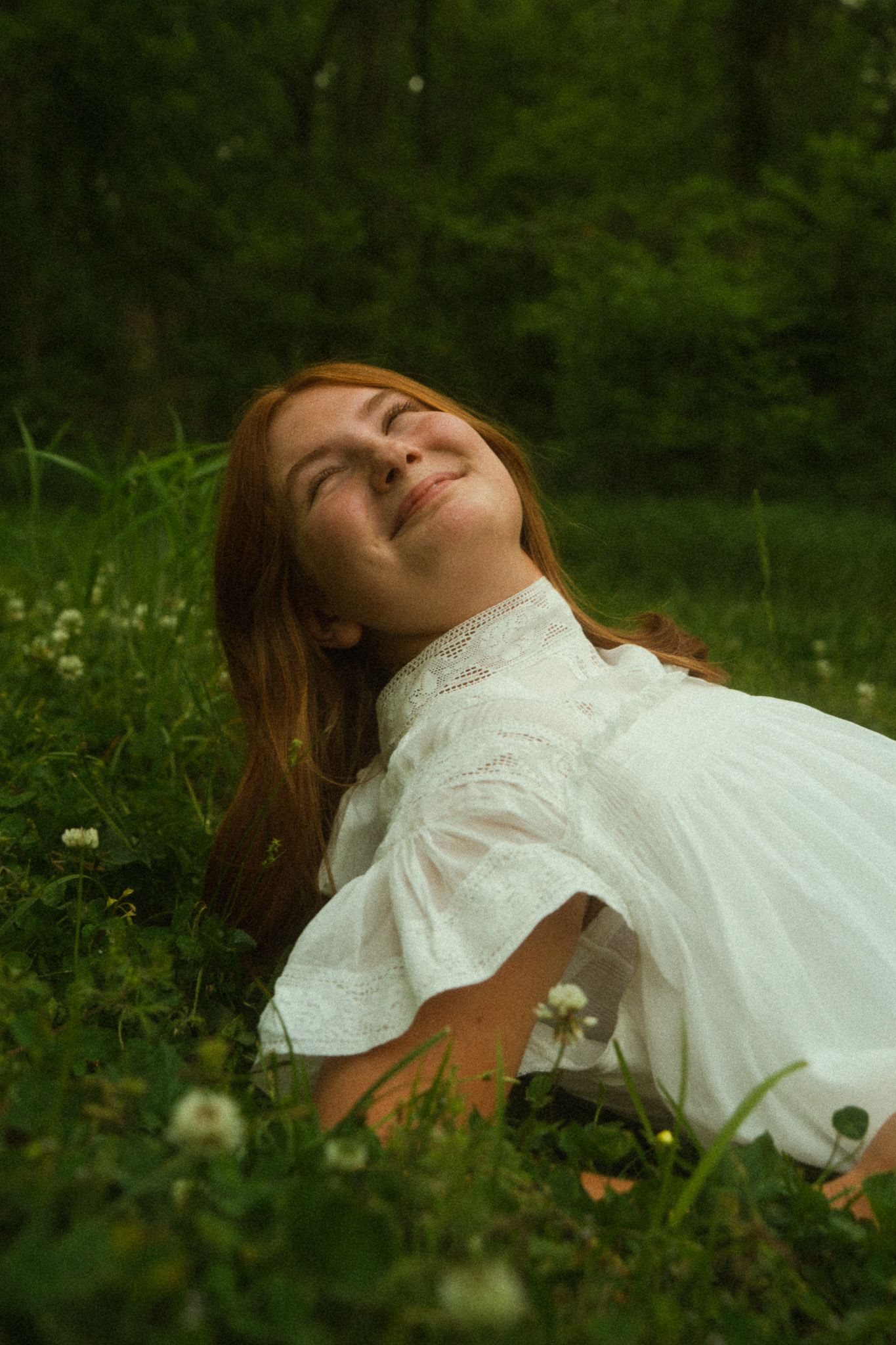 Young woman with red hair in white dress lying in green grass with wildflowers, smiling peacefully outdoors.