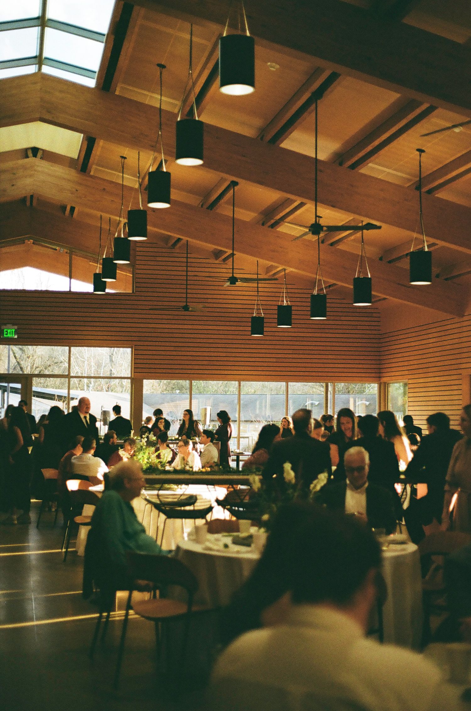 Large indoor reception venue with wooden beamed ceiling and pendant lighting filled with wedding guests.