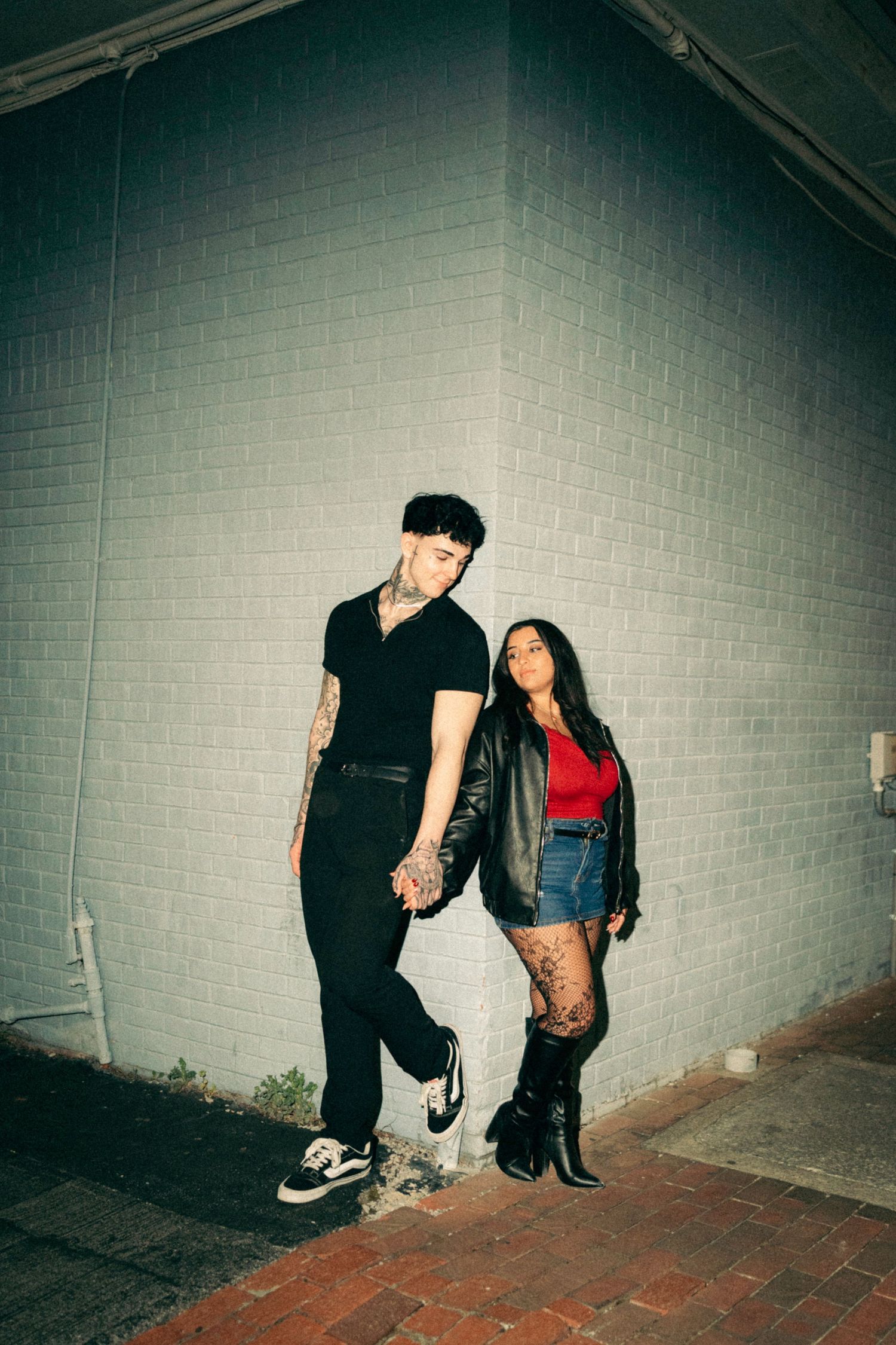Couple posing against brick wall at night, man in black outfit with tattoos, woman in red top, denim shorts, and boots.