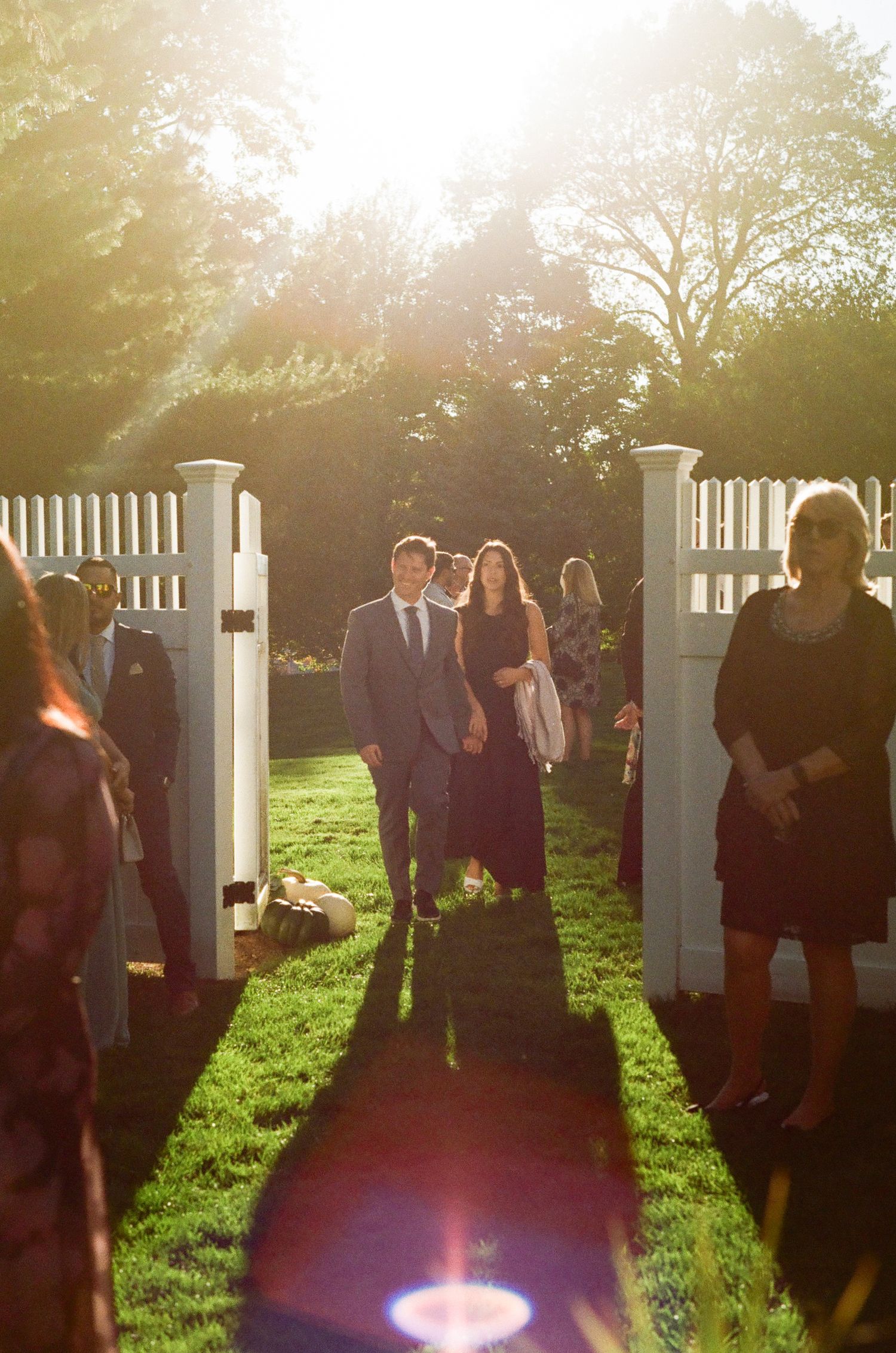 Sunlit garden entrance with white picket fence posts framing guests walking through.