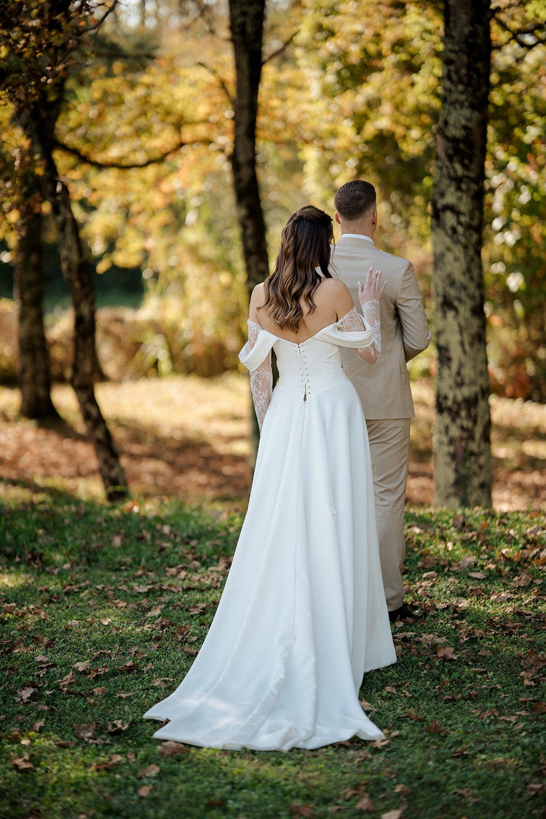 Bride in white off-shoulder gown and groom in beige suit walk hand-in-hand through autumn forest.