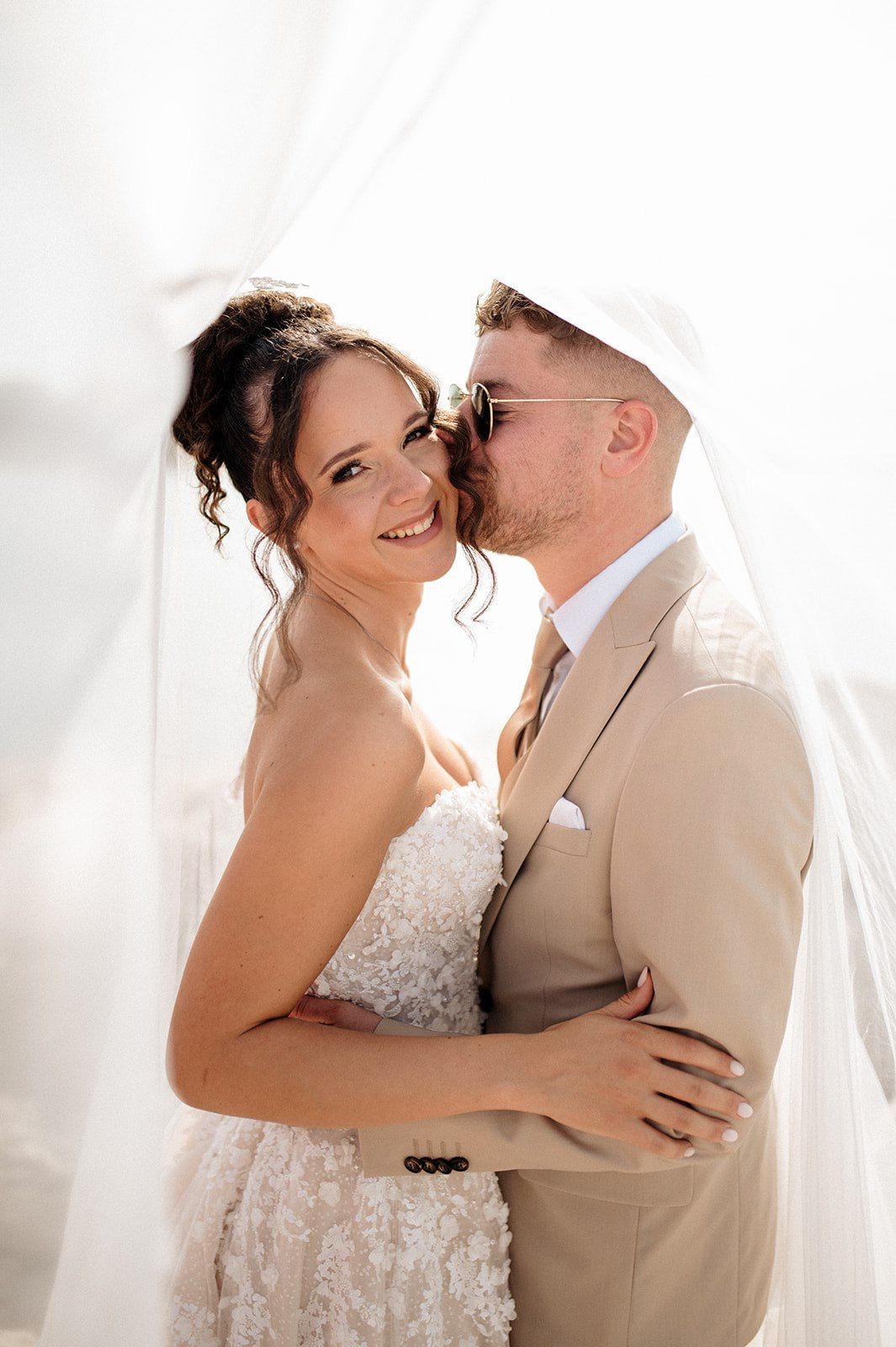 Bride and groom sharing a romantic kiss under a flowing veil on their wedding day.