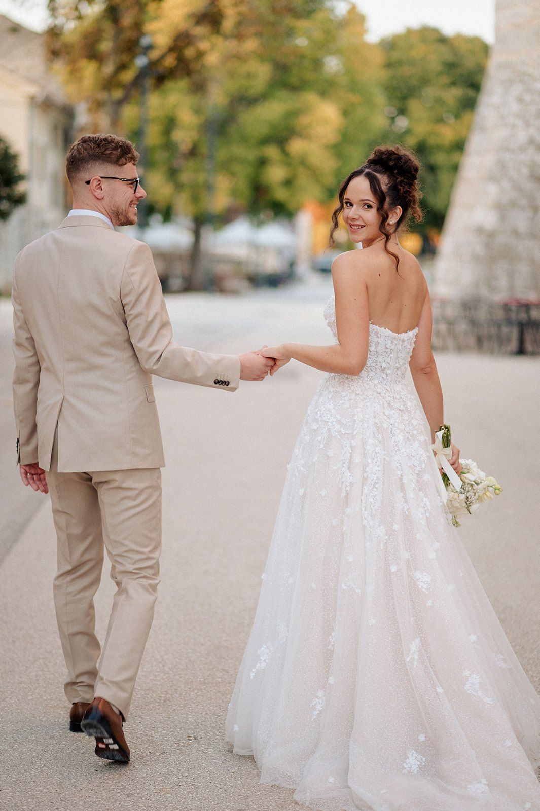 A bride in a floral lace gown holds hands with a groom in a beige suit as they walk outdoors on their wedding day.