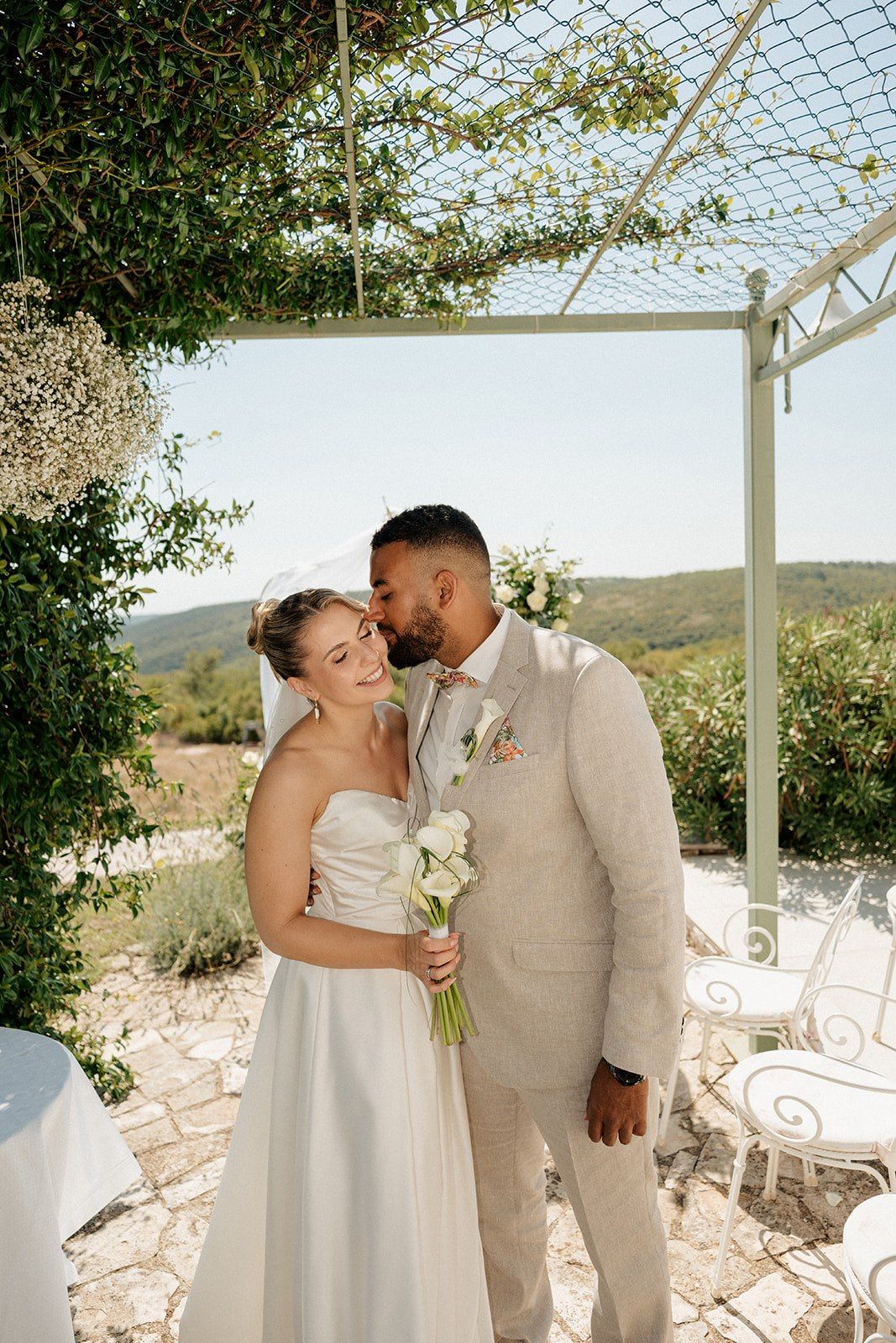 Bride and groom share a romantic moment under a pergola, with lush greenery and scenic hills in the background.