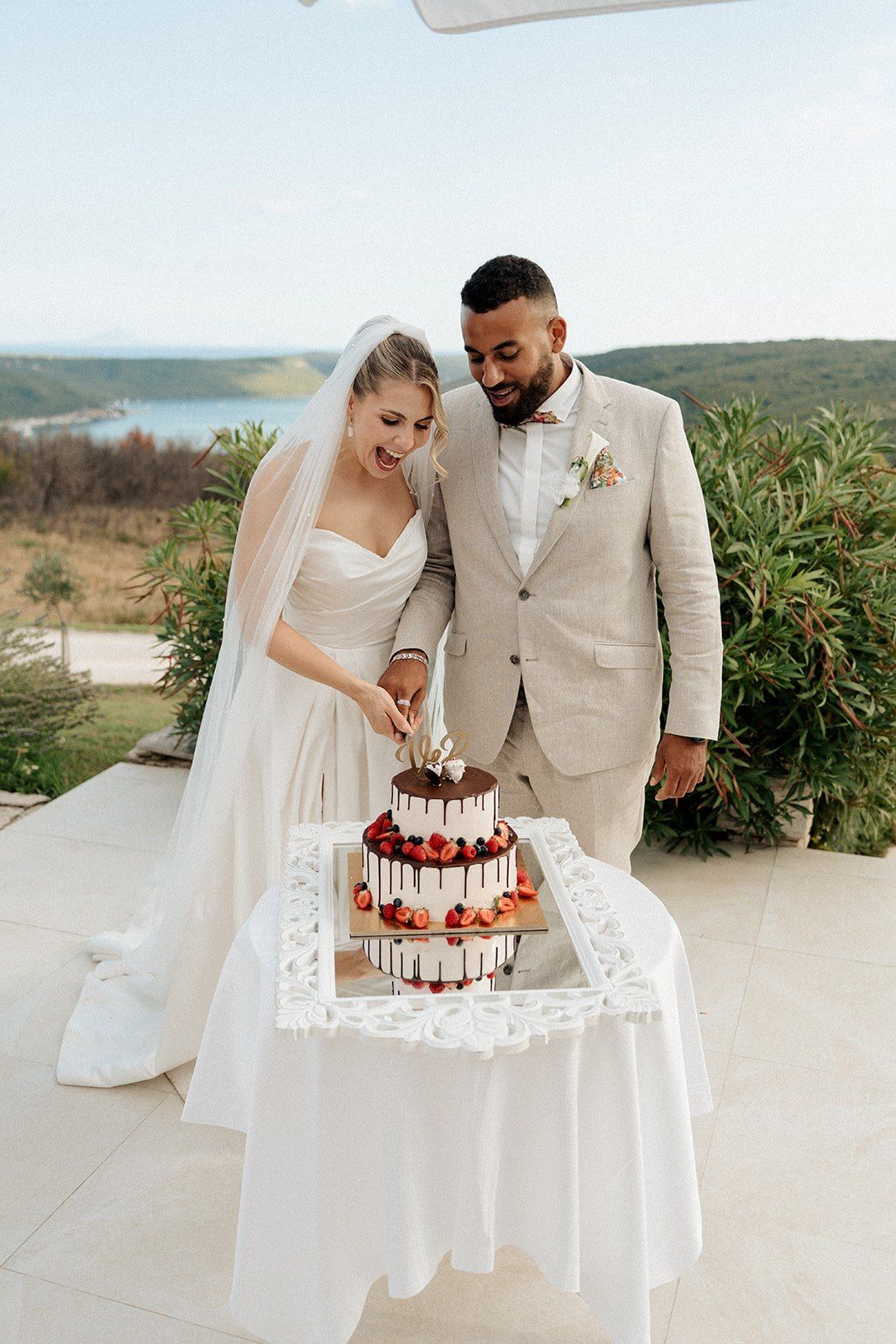 Bride and groom cut a three-tier chocolate drip wedding cake decorated with strawberries at outdoor reception.