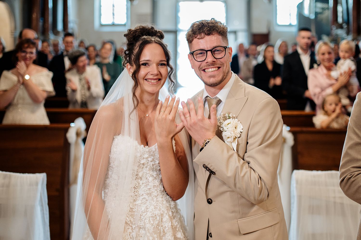 Newlywed couple smiling and showing wedding rings in church aisle, bride in white gown with veil, groom in beige suit.