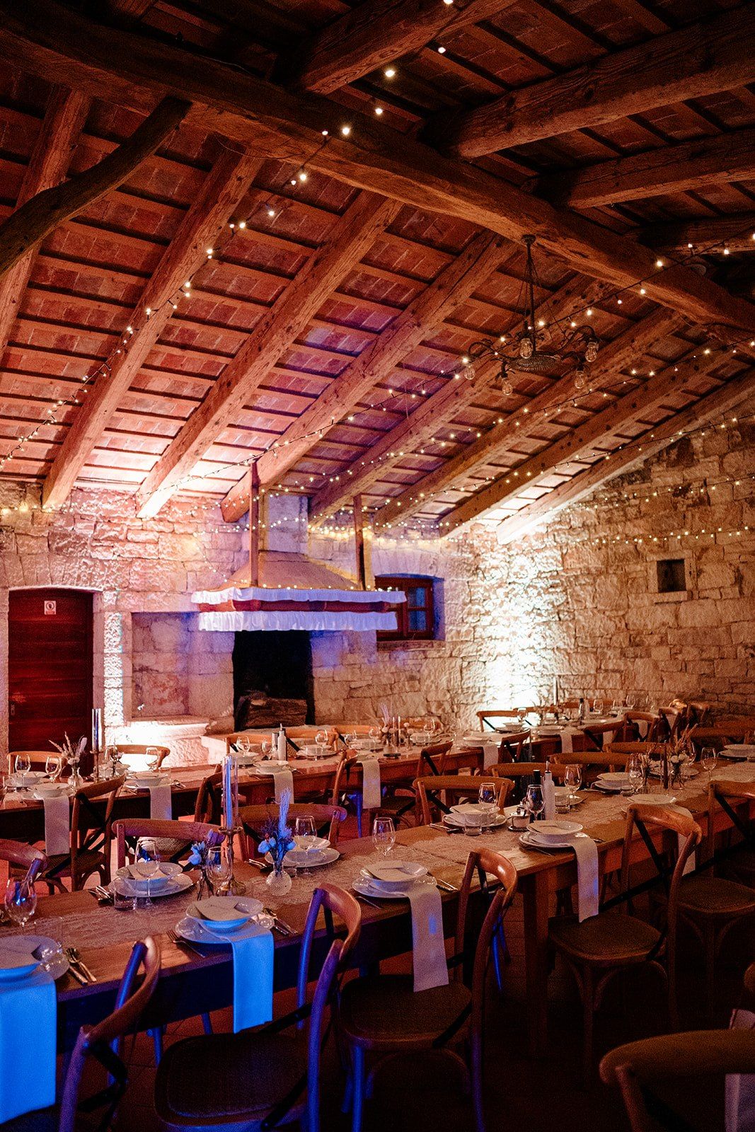 Rustic wedding reception in a stone venue with wooden beam ceiling, fairy lights, and long dining tables set for guests.
