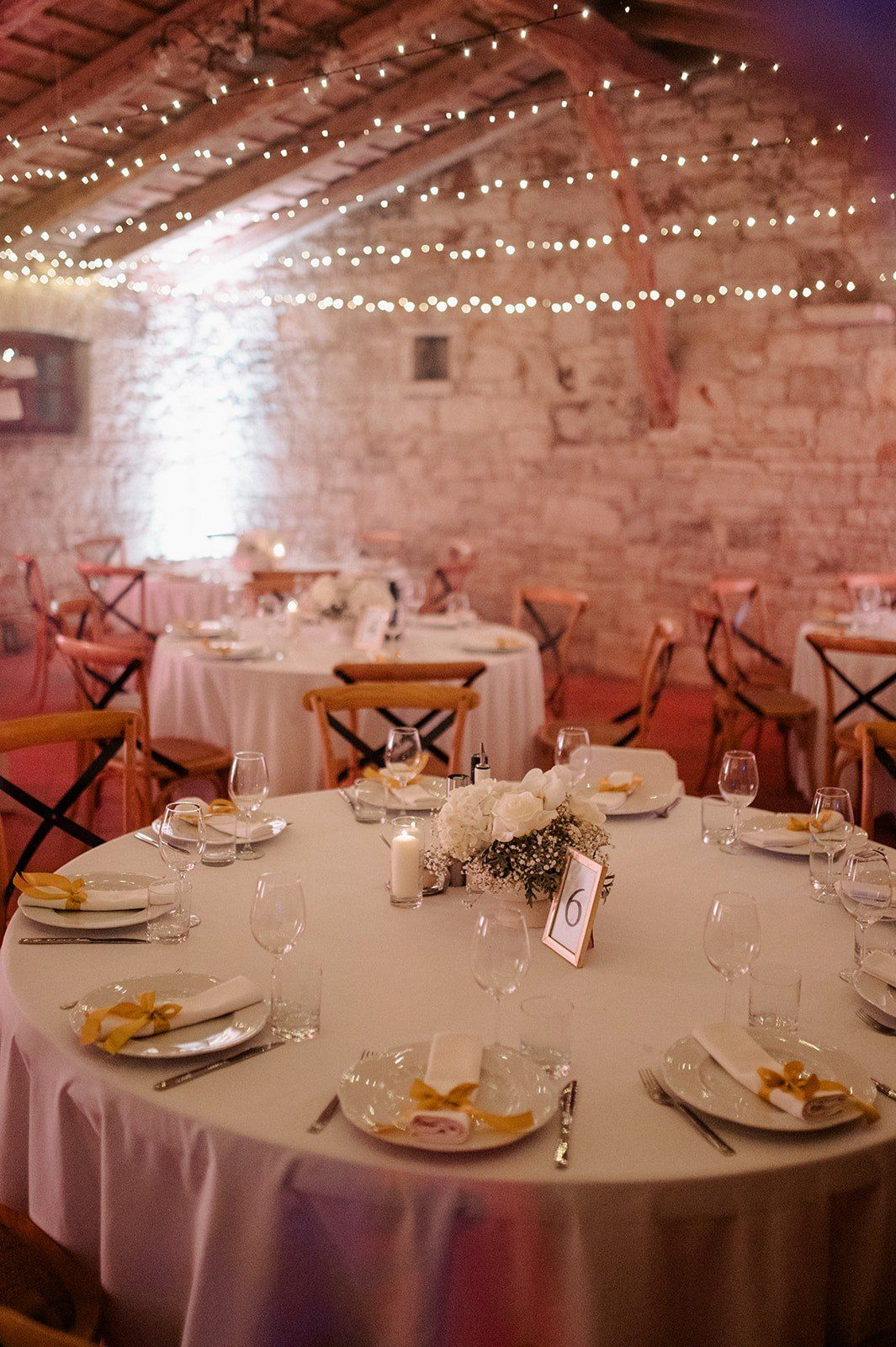 Elegant barn wedding reception with fairy lights, round tables, white linens, floral centerpieces, and rustic stone walls.