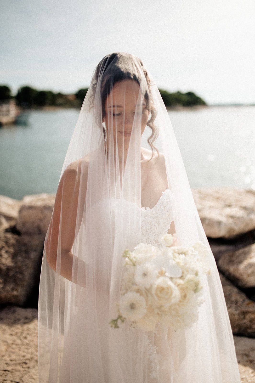 Bride in white lace gown and flowing veil holds white floral bouquet by rocky waterfront on wedding day.