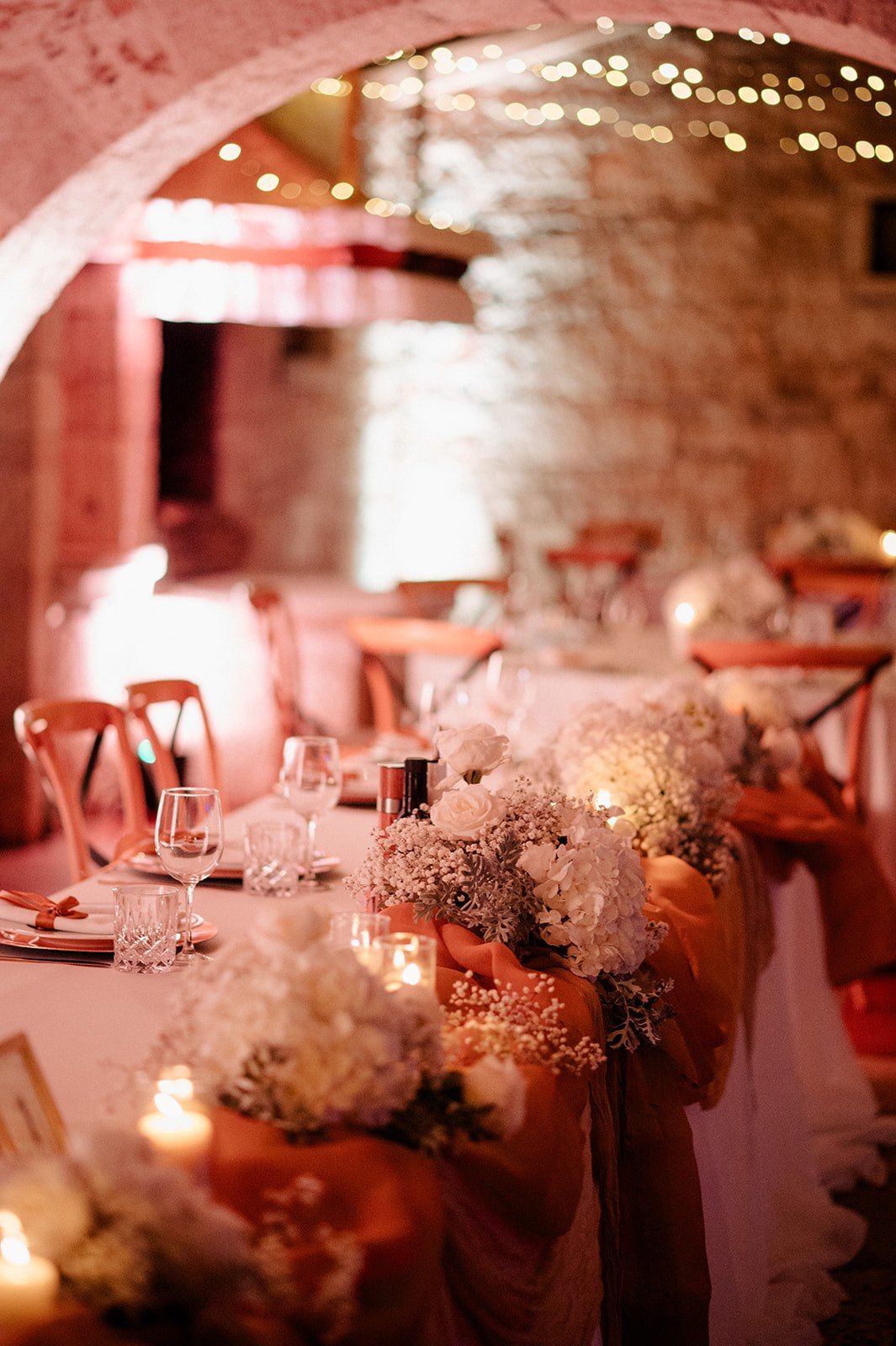 Elegant wedding reception in a stone venue with candlelit tables, white floral centerpieces, and warm pink ambient lighting.