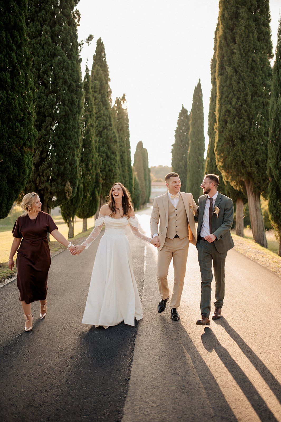 Bride and groom walk hand-in-hand with guests down a cypress-lined road in golden hour sunlight.
