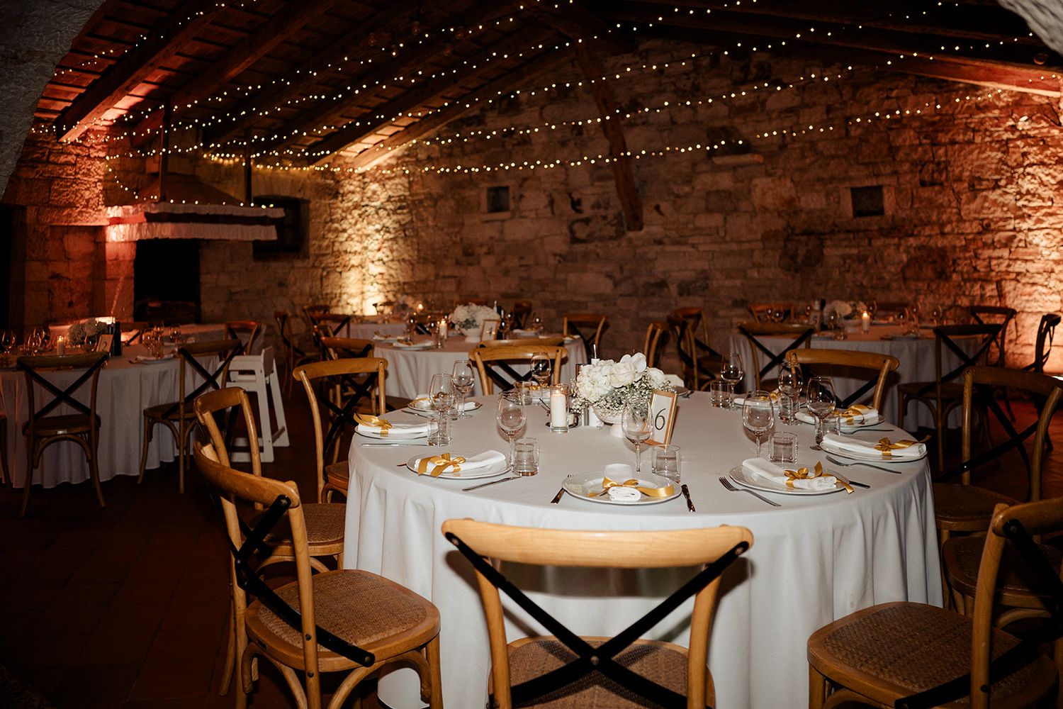 Elegant wedding reception in a rustic stone venue with fairy lights, round tables, white linens, and wooden cross-back chairs.