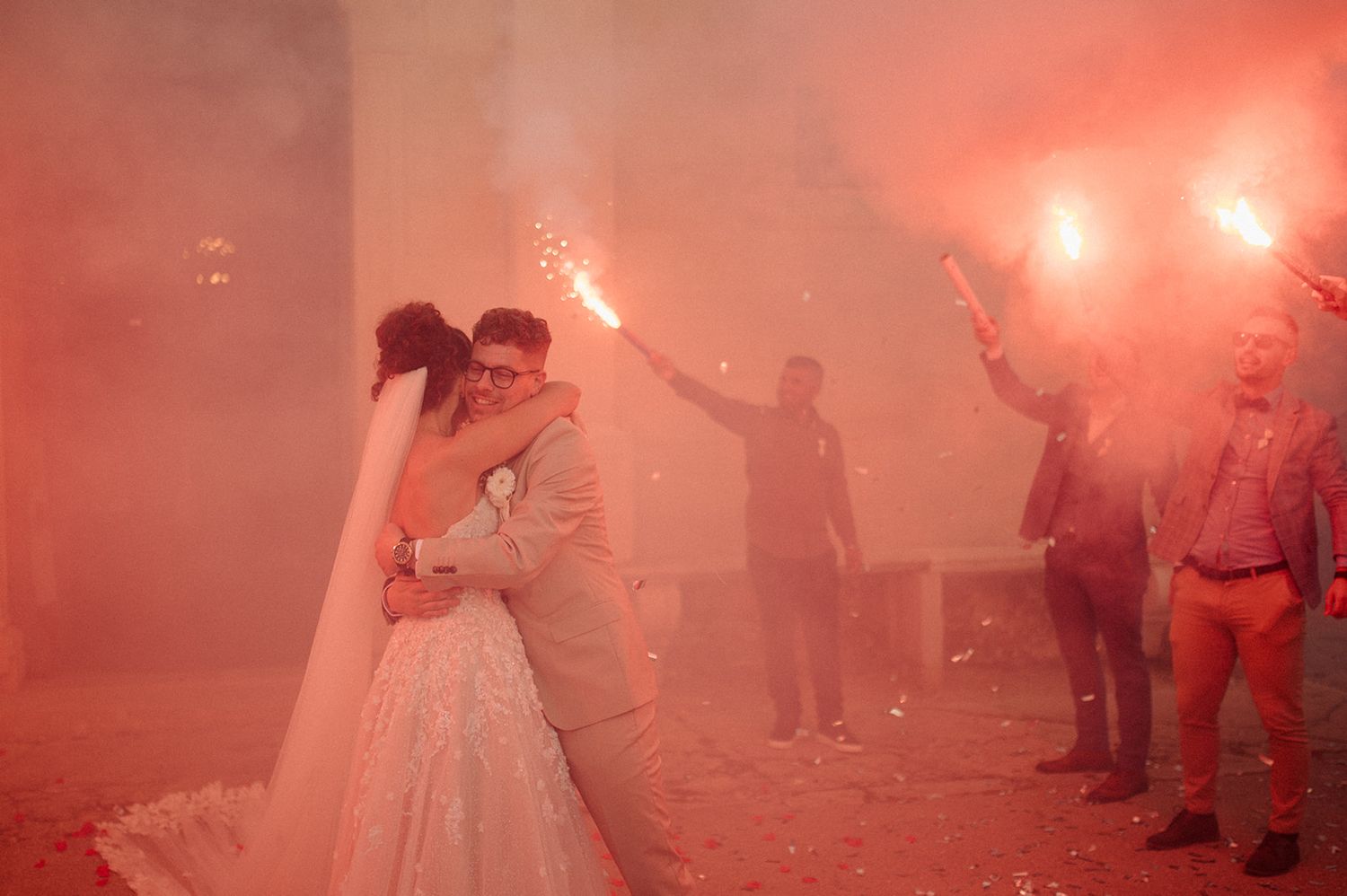Bride and groom kissing surrounded by guests holding red smoke flares at an outdoor wedding celebration.