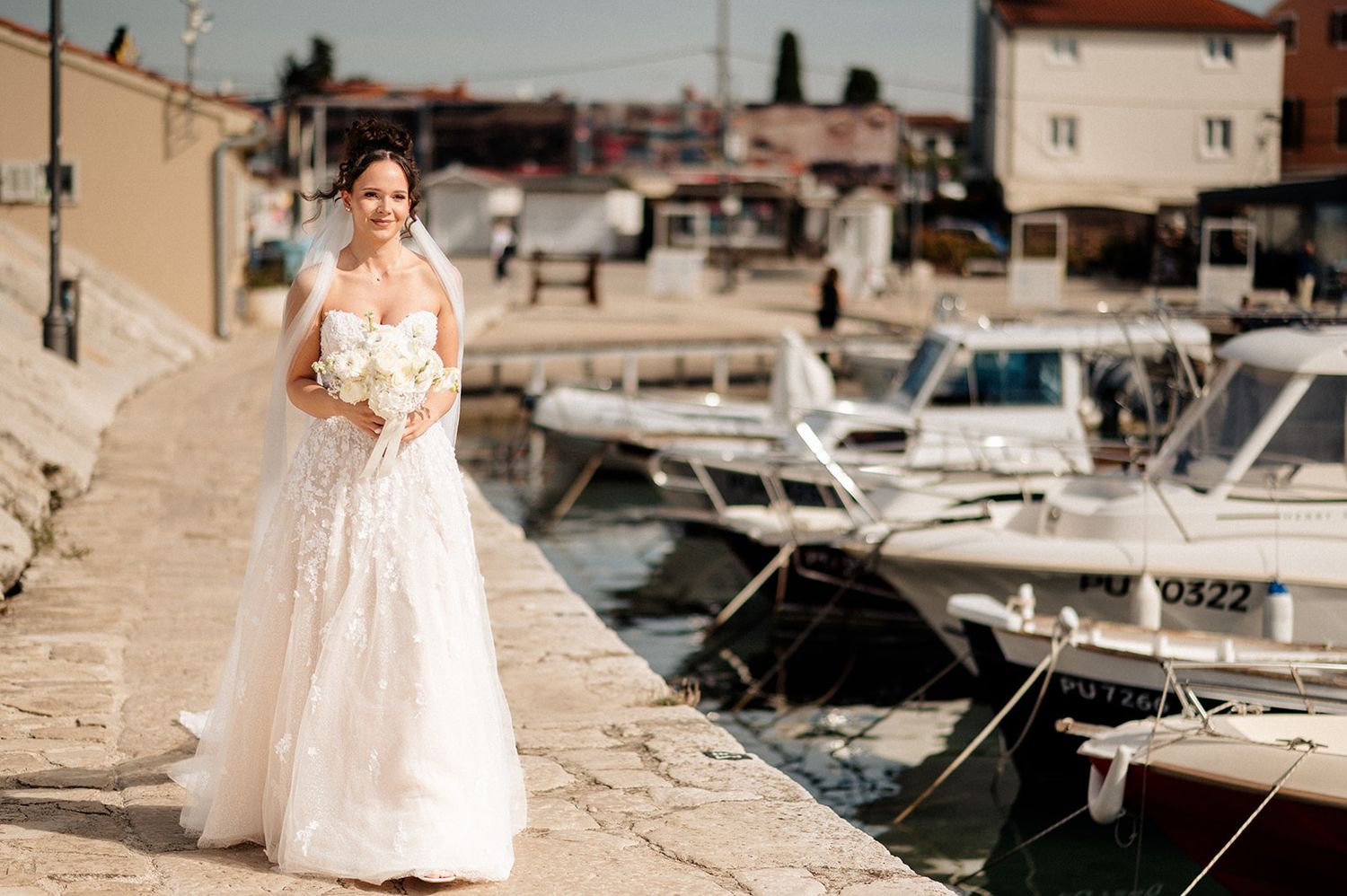 Bride in white lace gown holding bouquet walks along marina with boats docked in background on sunny day.