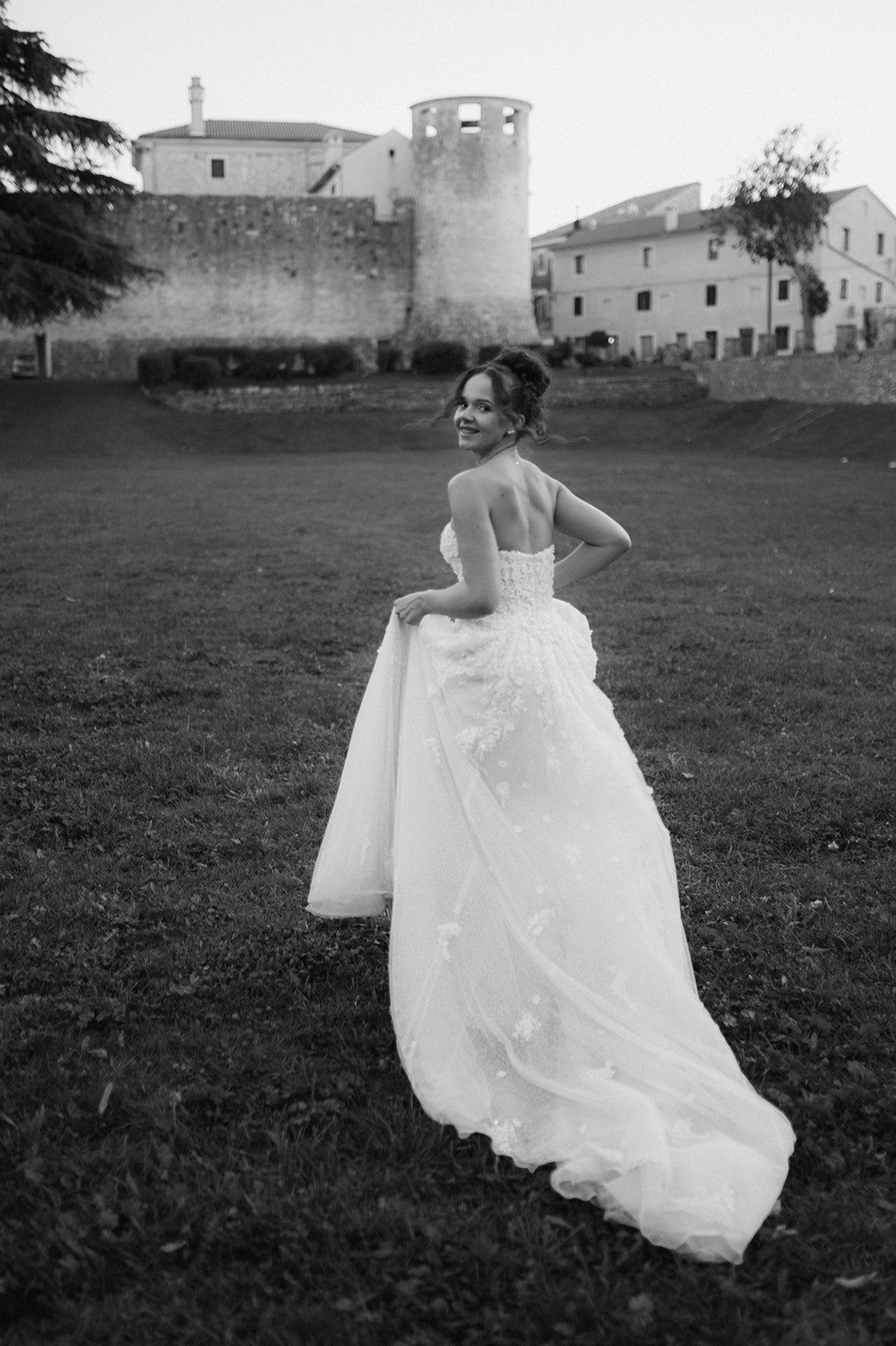 Bride in a white ball gown with train smiles over her shoulder in a black and white photo near a historic stone castle.