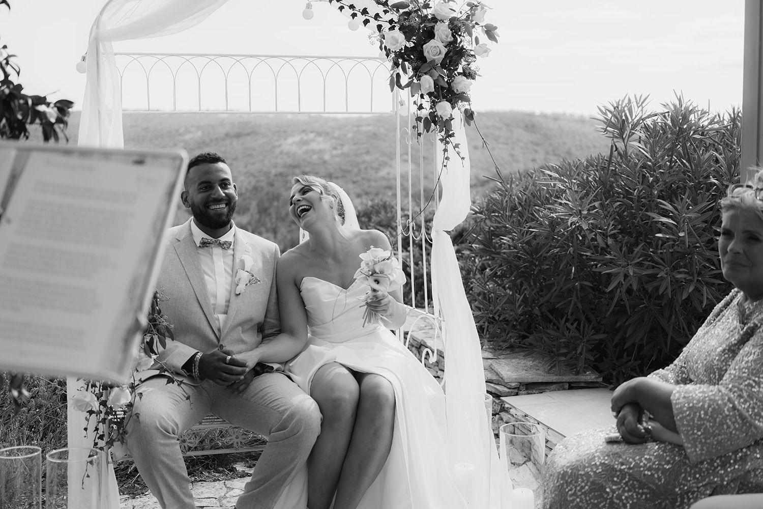 Black and white photo of smiling bride and groom seated together during outdoor wedding ceremony.