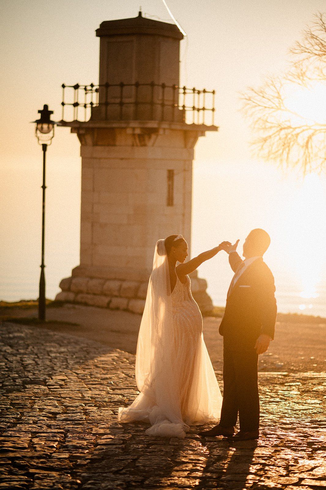 A romantic sunset wedding photo in front of a historic lighthouse with cobblestone ground.